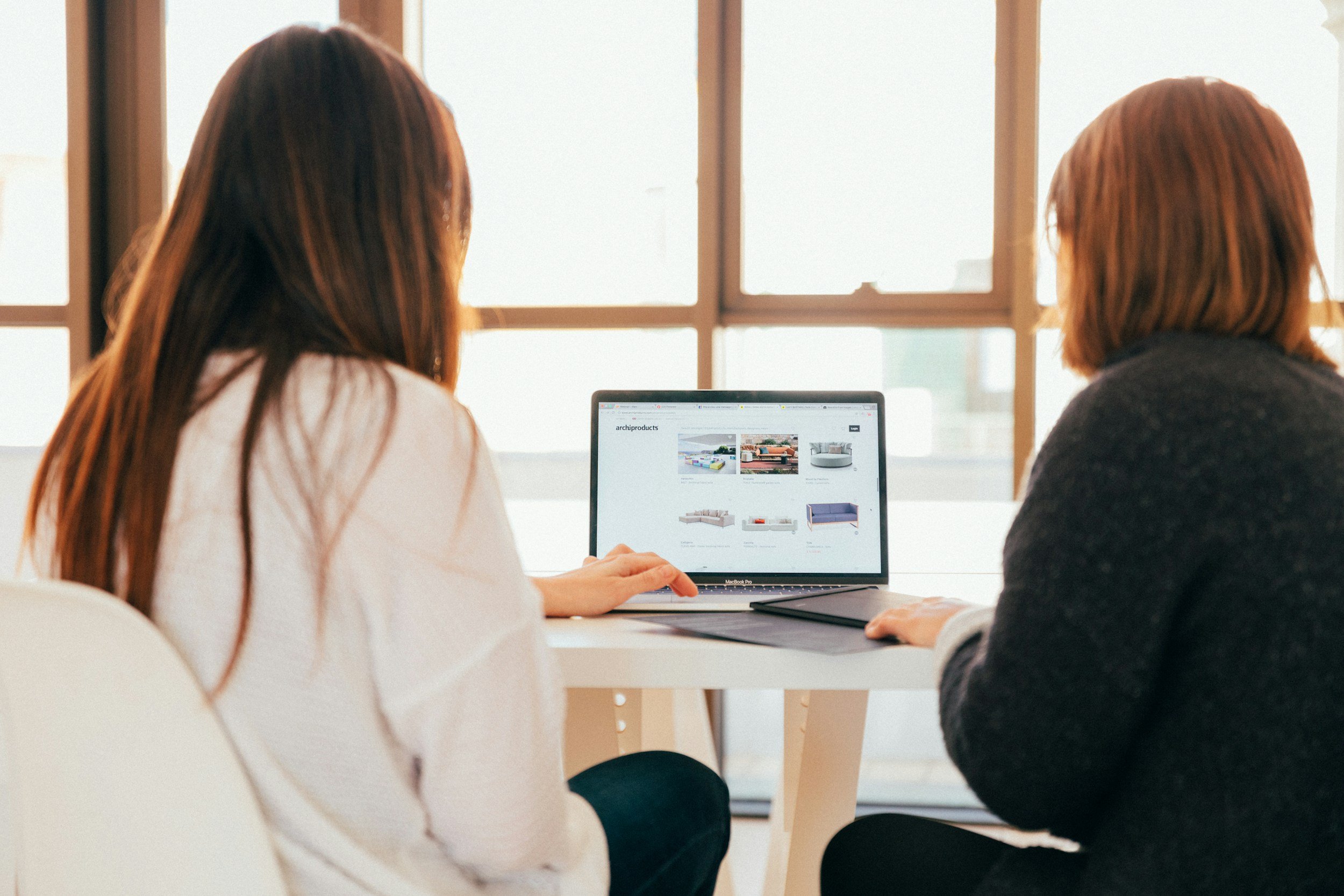 Two women sitting at a table looking at a laptop screen showing a website with furniture images.
