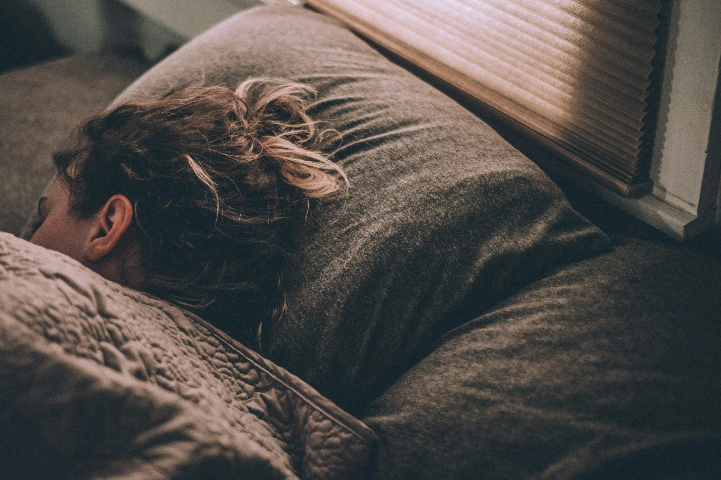 A person with wavy hair lying on a couch, resting with eyes closed, covered with a textured blanket, next to a window with closed blinds.