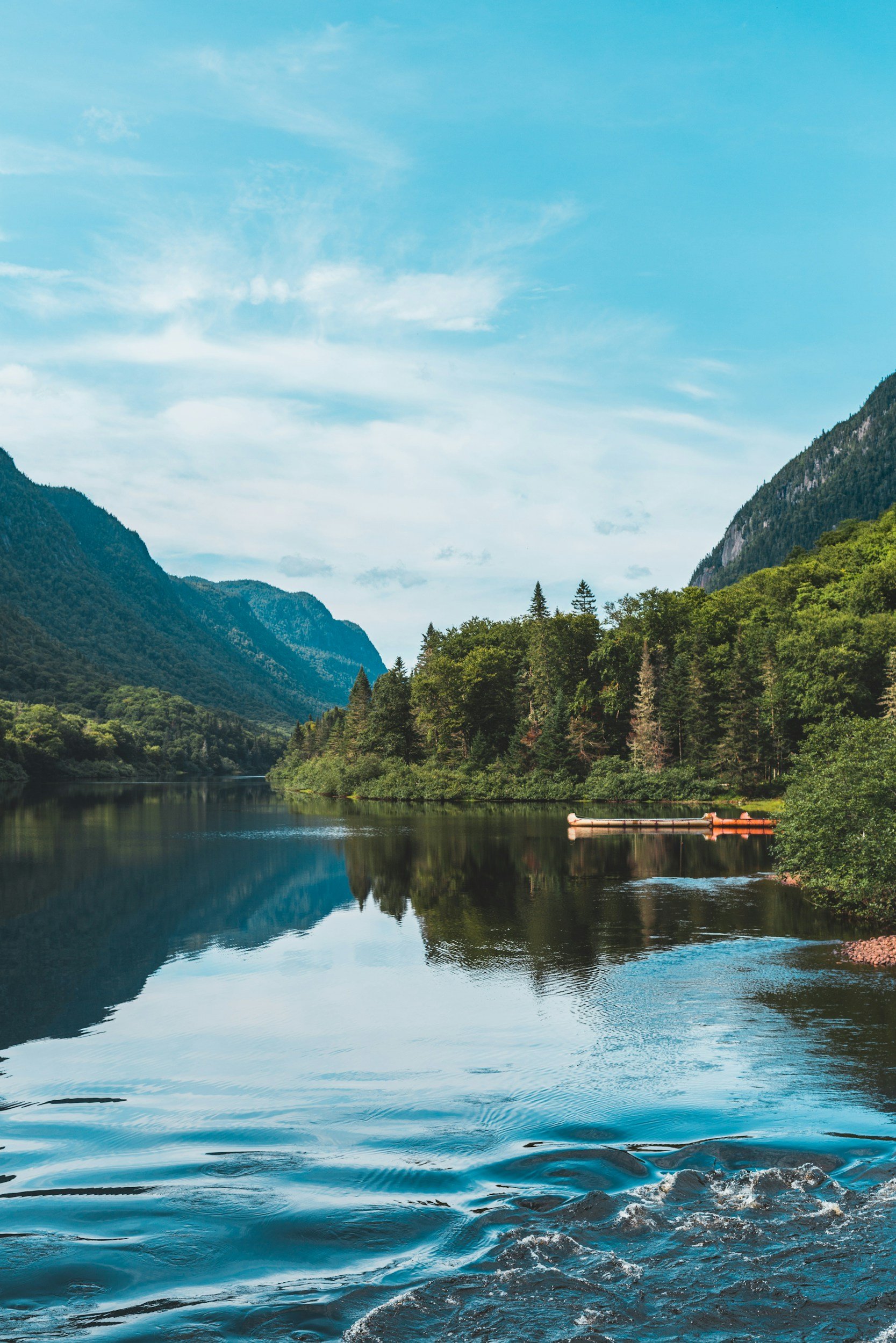 Scenic view of a calm river surrounded by lush green forests and mountains under a partly cloudy sky.