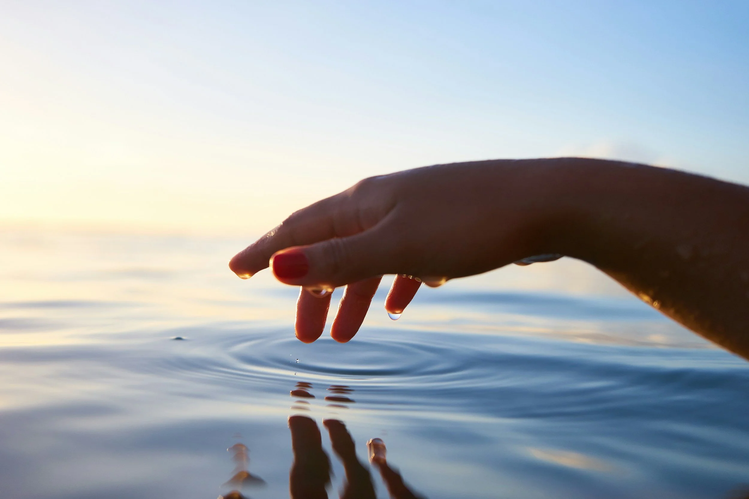 A hand reaches out over calm water during a sunset, with the sky transitioning from light to darker blue.