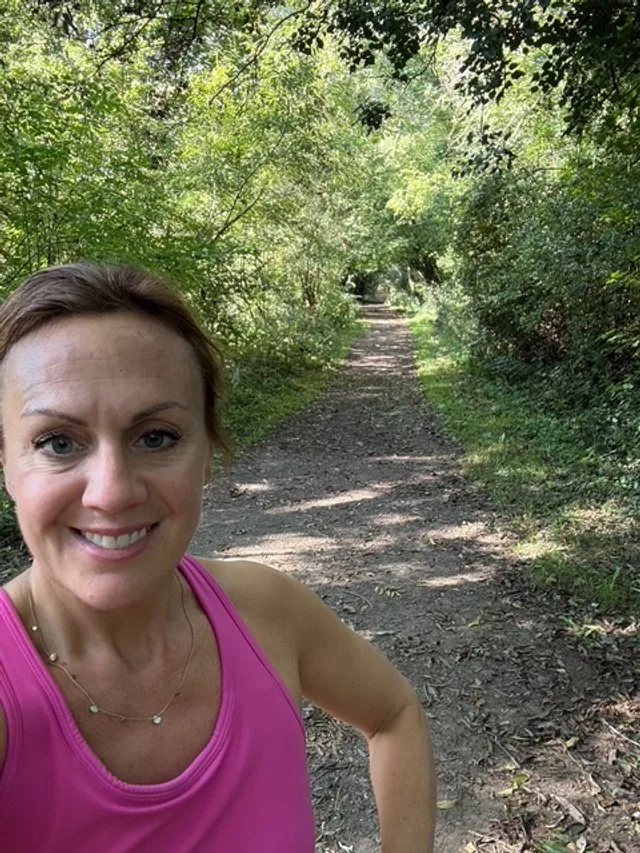 A woman in a pink tank top taking a selfie on a dirt trail surrounded by lush green trees.