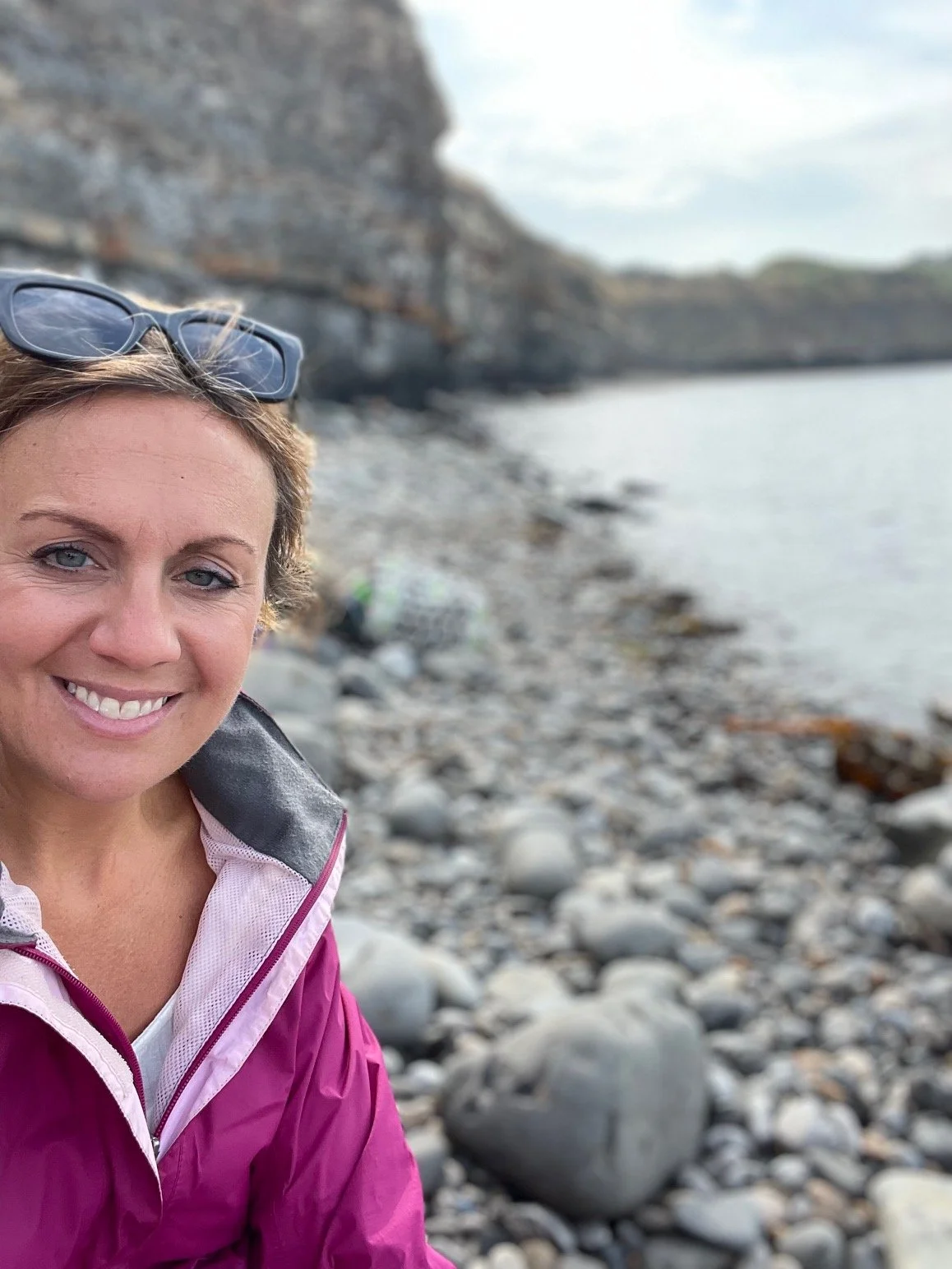 Smiling woman with short hair wearing glasses on her head and a pink jacket, sitting on a rocky beach with a cliff and water in the background.