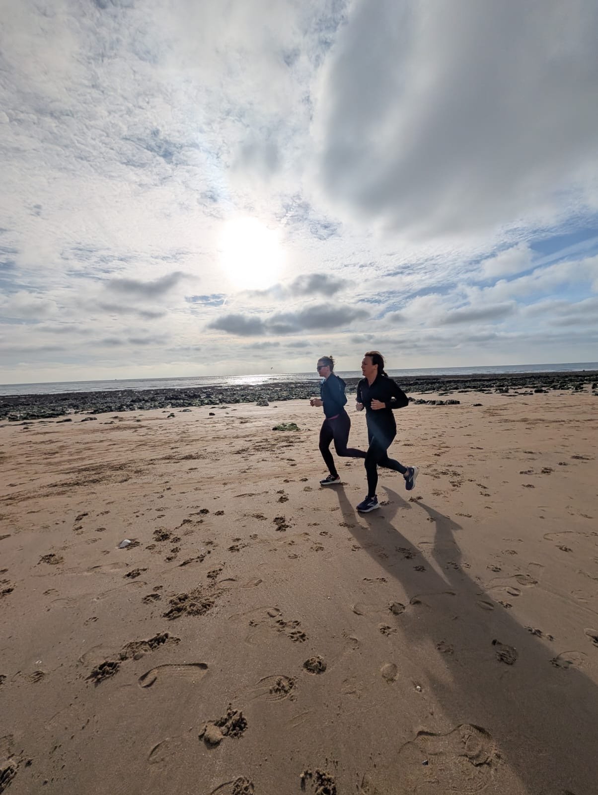 Two women jogging on a sandy beach under a cloudy sky with the sun partially obscured, footprints visible in the sand.
