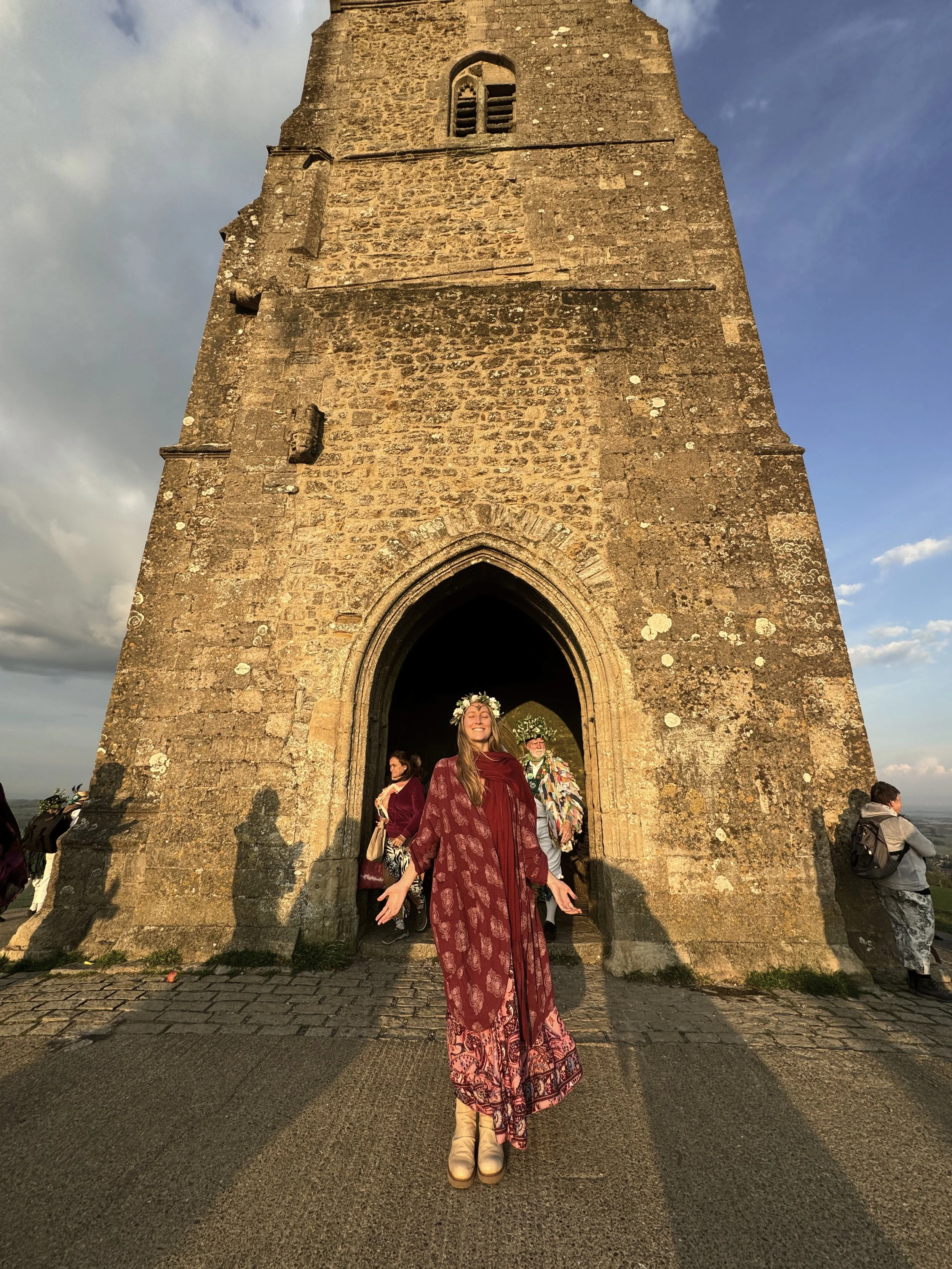 Una mujer feliz con vestido largo y una corona de flores en la cabeza, de pie frente a una estructura antigua de piedra, en un acontecimiento que parece un festival o ceremonia en la tarde.