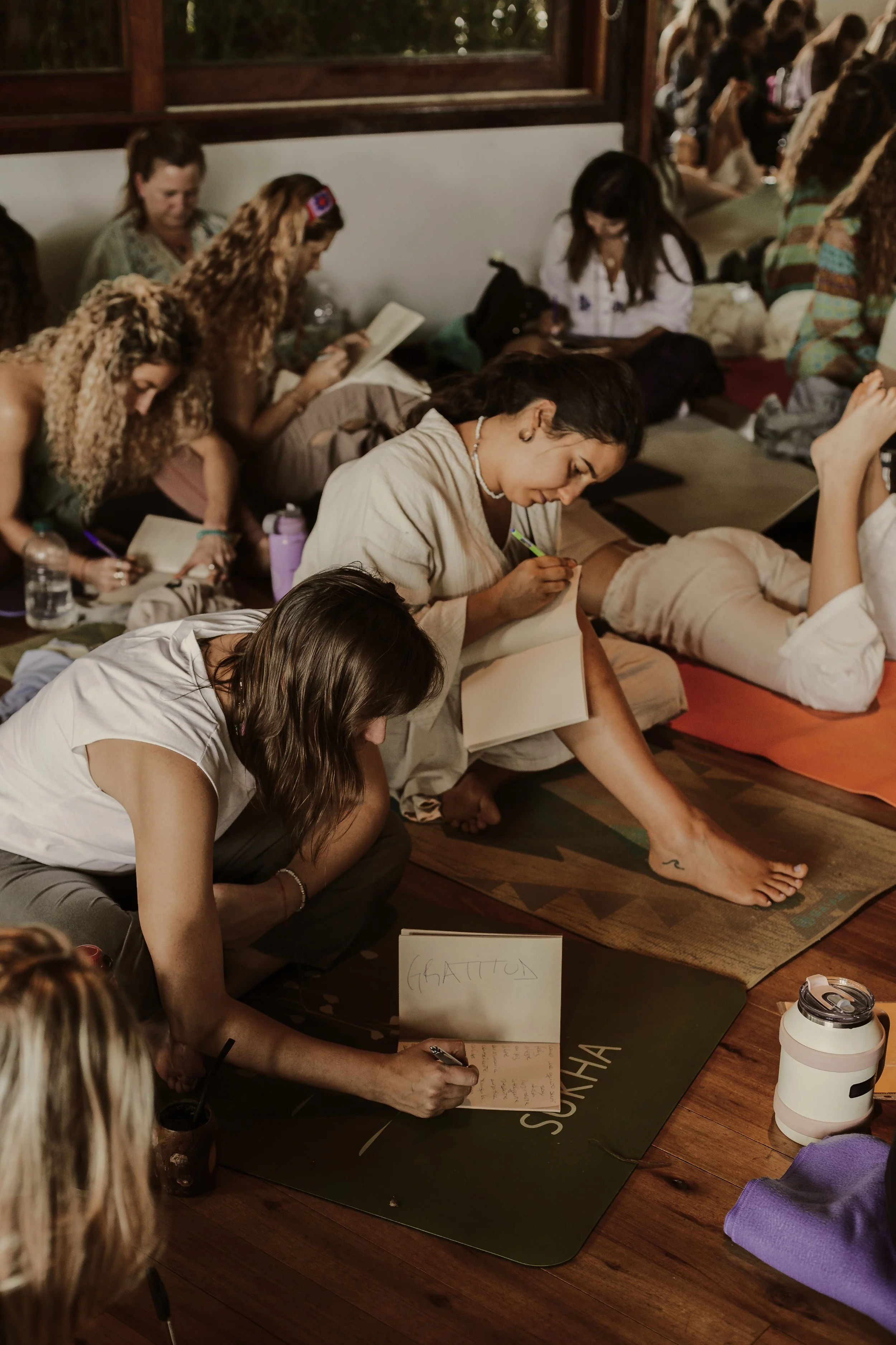 Grupo de personas en un taller, la mayoría sentadas en el suelo con libretas y libros, escribiendo o leyendo en un ambiente interior con paredes de madera y ventanas.