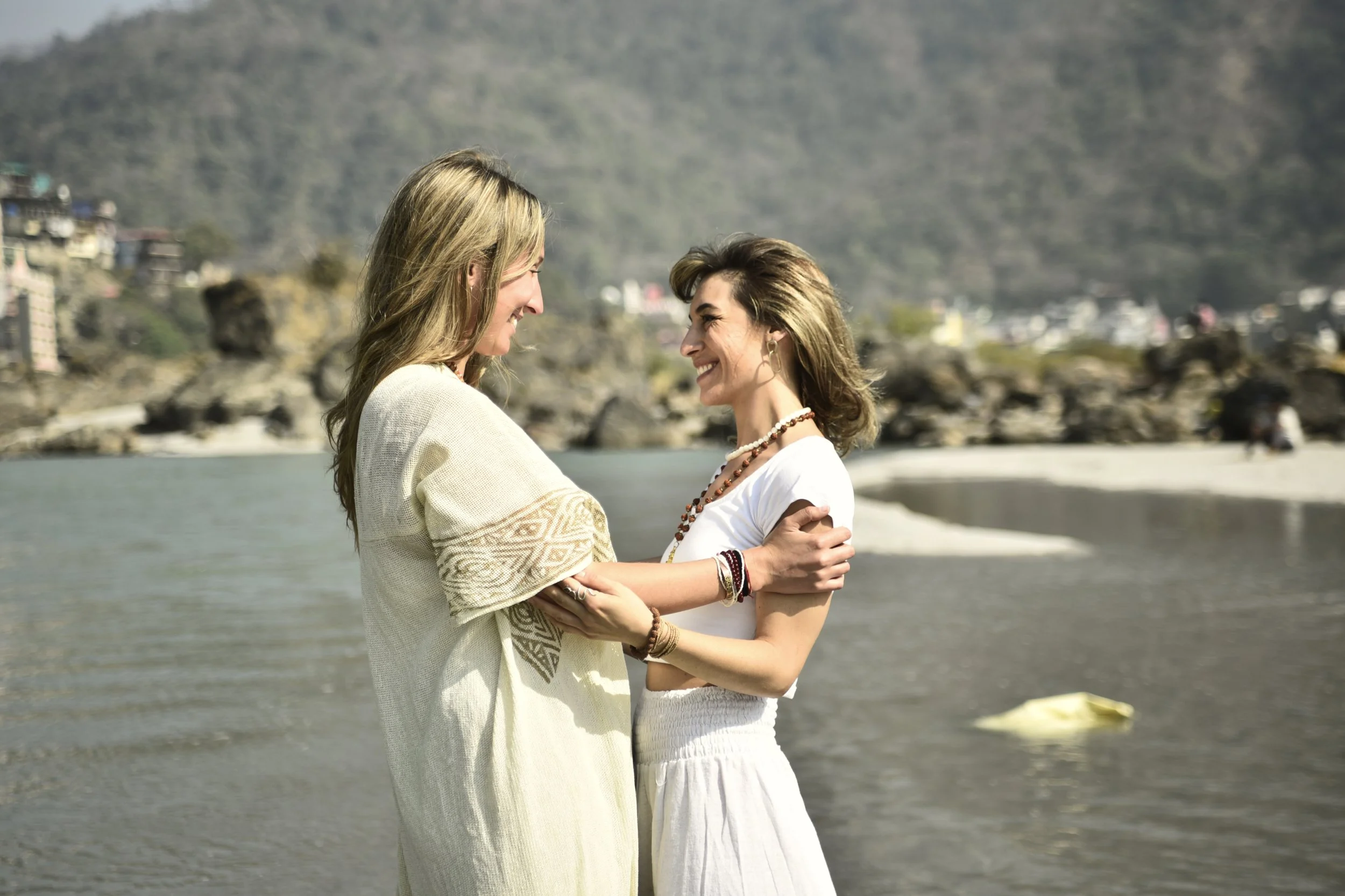 Dos mujeres sonriendo en la orilla de un río o lago rodeadas de montañas y rocas, en un ambiente natural y tranquilo.