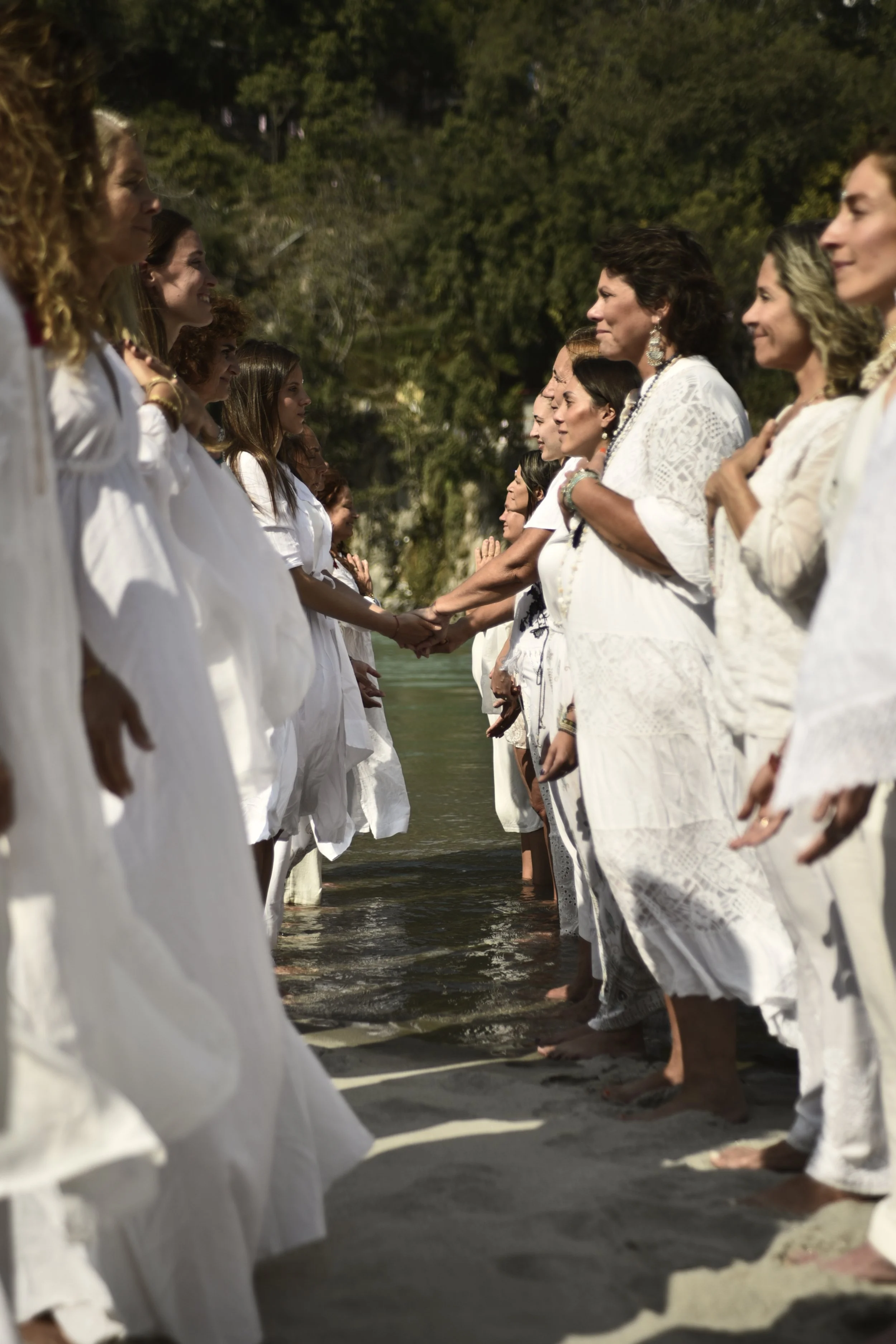 Mujeres con vestidos blancos de ceremonia en una ceremonia en la orilla del río, tomadas de las manos.
