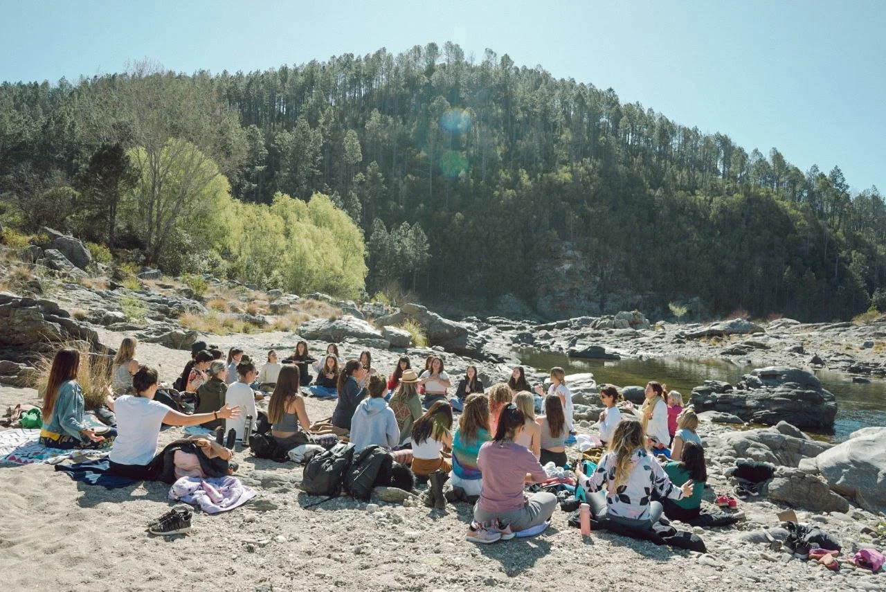 Grupo de personas sentadas en rocas junto a un río en un día soleado, rodeados de árboles y montañas.