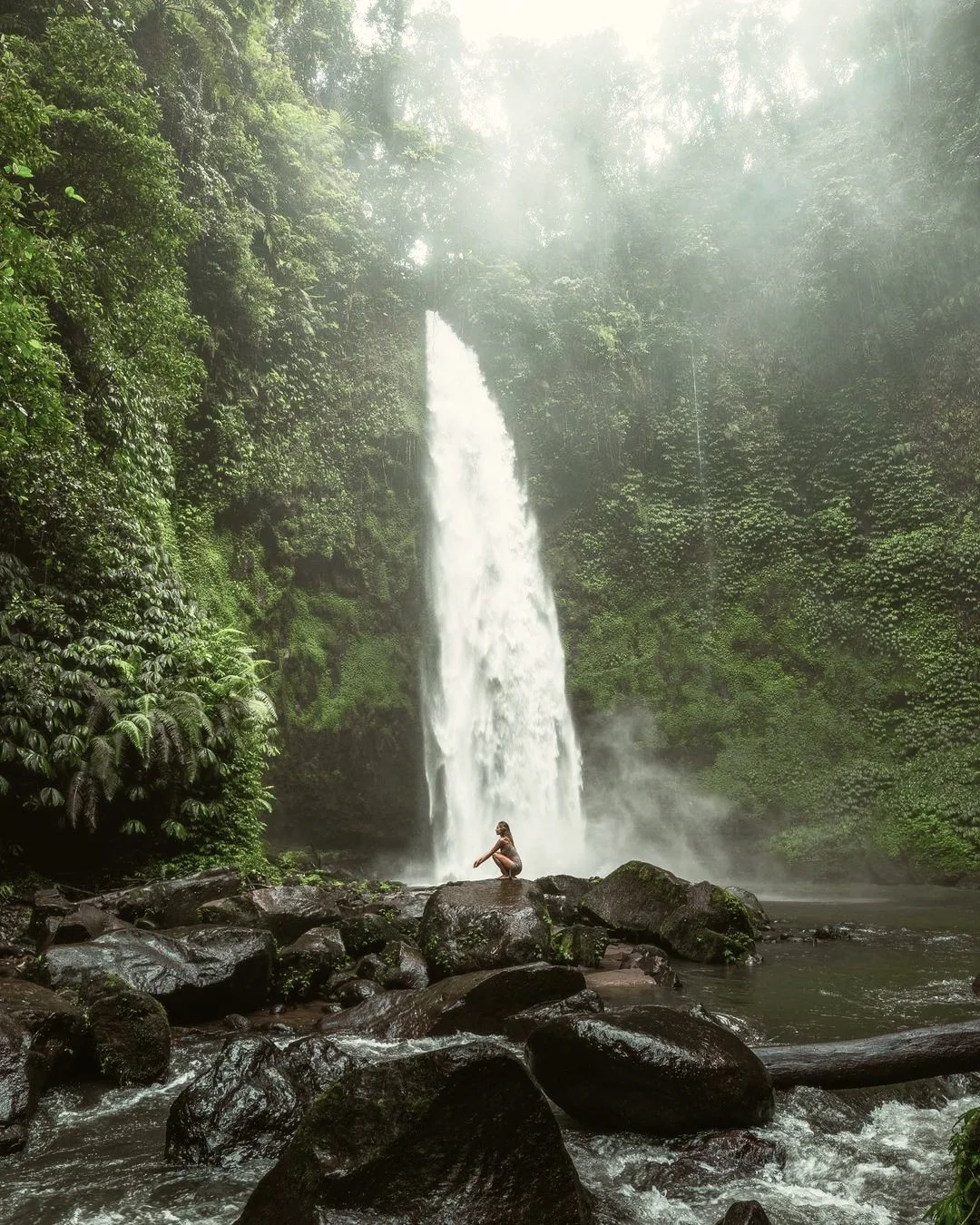 Una cascada en medio de un bosque verde, con una persona sentada en rocas frente a la cascada, rodeada de vegetación y rocío.