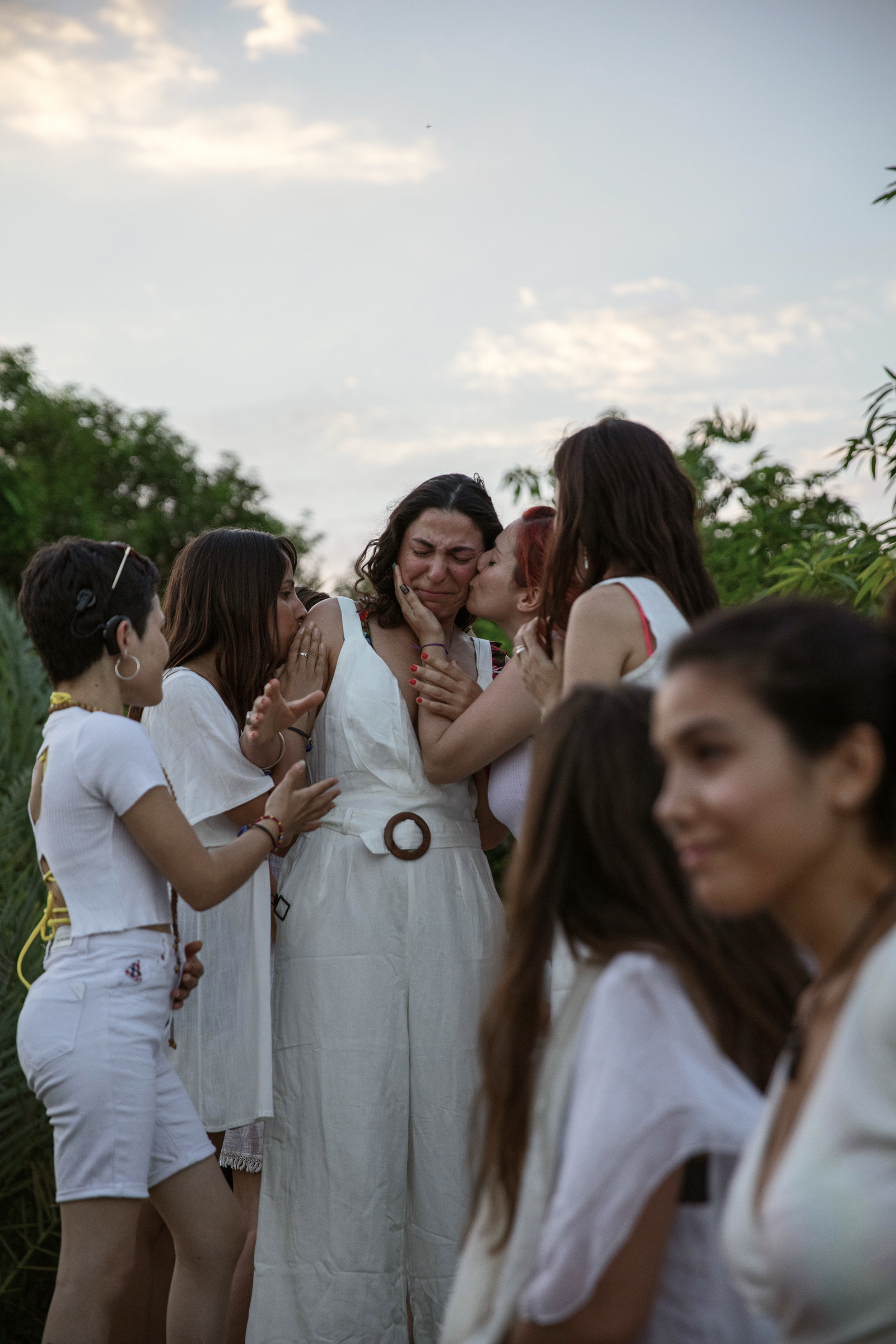 Grupo de mujeres y niñas en una reunión emotiva al aire libre, algunas acurrucadas, en un día soleado con cielo parcialmente nublado.