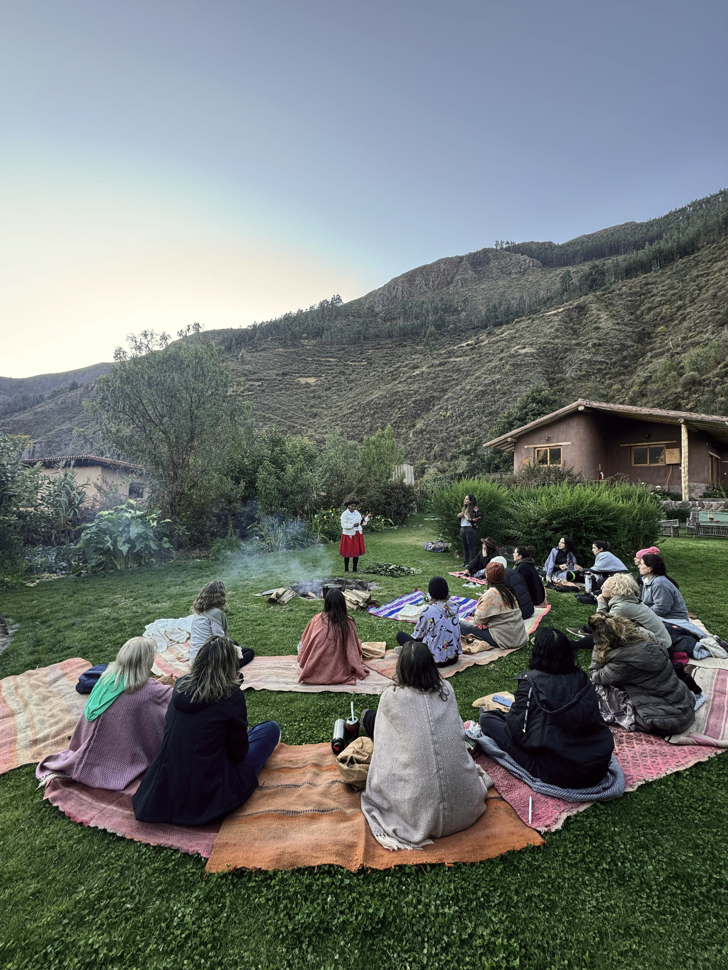 Un grupo de personas sentadas en alfombrillas de diferentes colores en un campo de césped, participando en una ceremonia o evento al aire libre, mientras una mujer de pie en el centro parece hablar o presentar. Al fondo, montañas y casas.