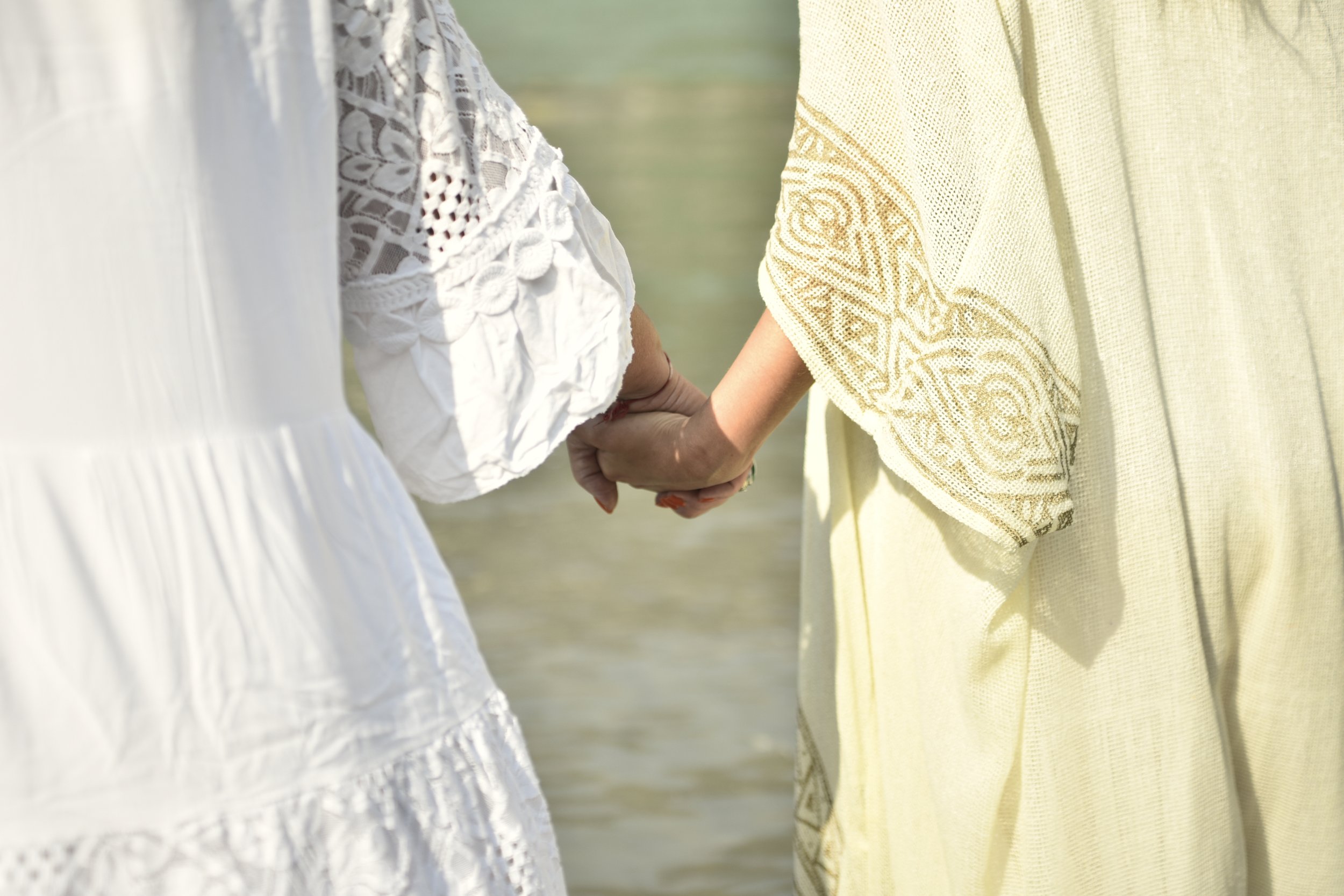 Dos personas de vestimenta clara sosteniendo sus manos unidas, vista de espalda y en un entorno natural, cerca del agua.