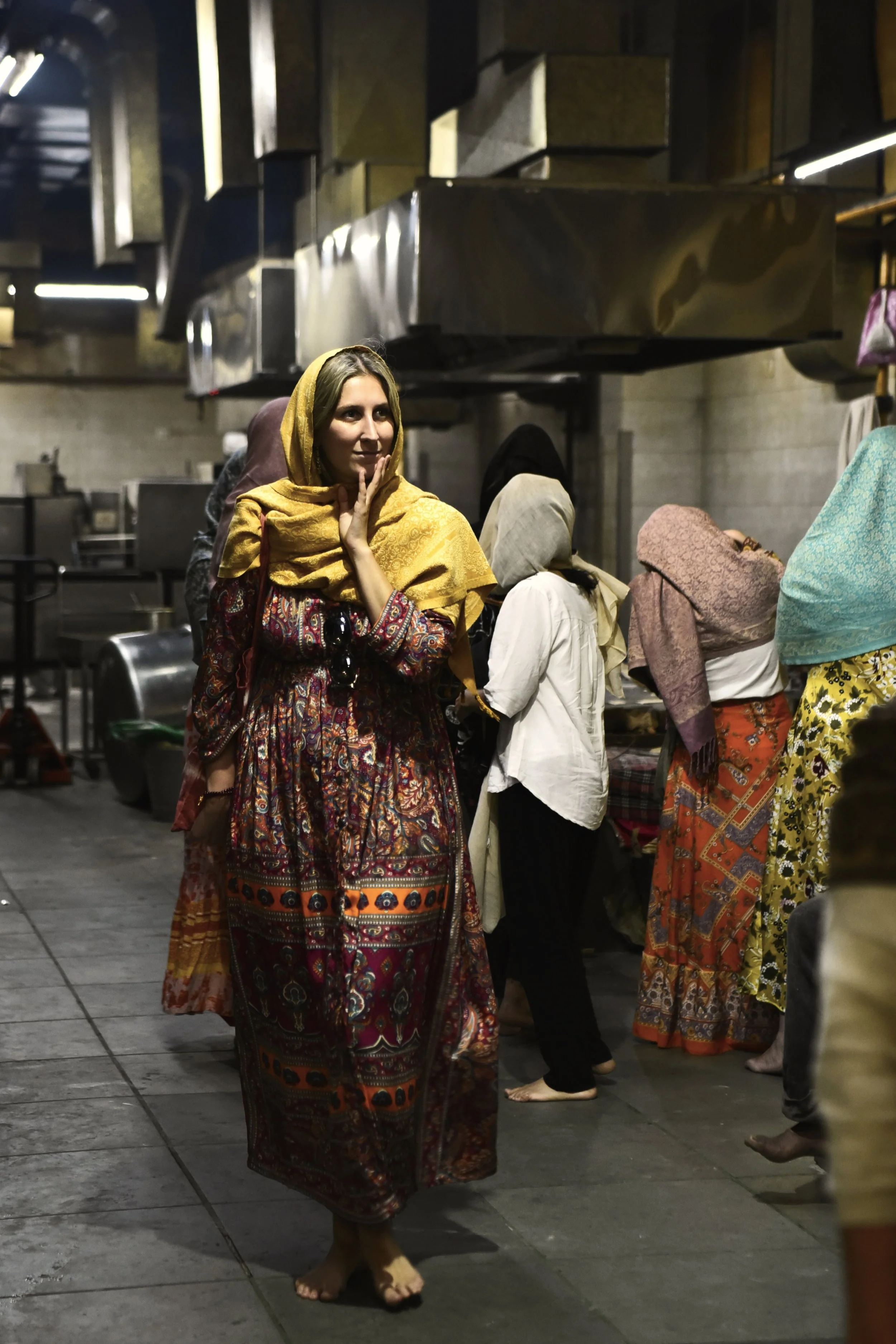 Mujer vestida con ropas tradicionales con un pañuelo amarillo en la cabeza, en un lugar con varias mujeres con vestimenta similar y cocinas de fondo.