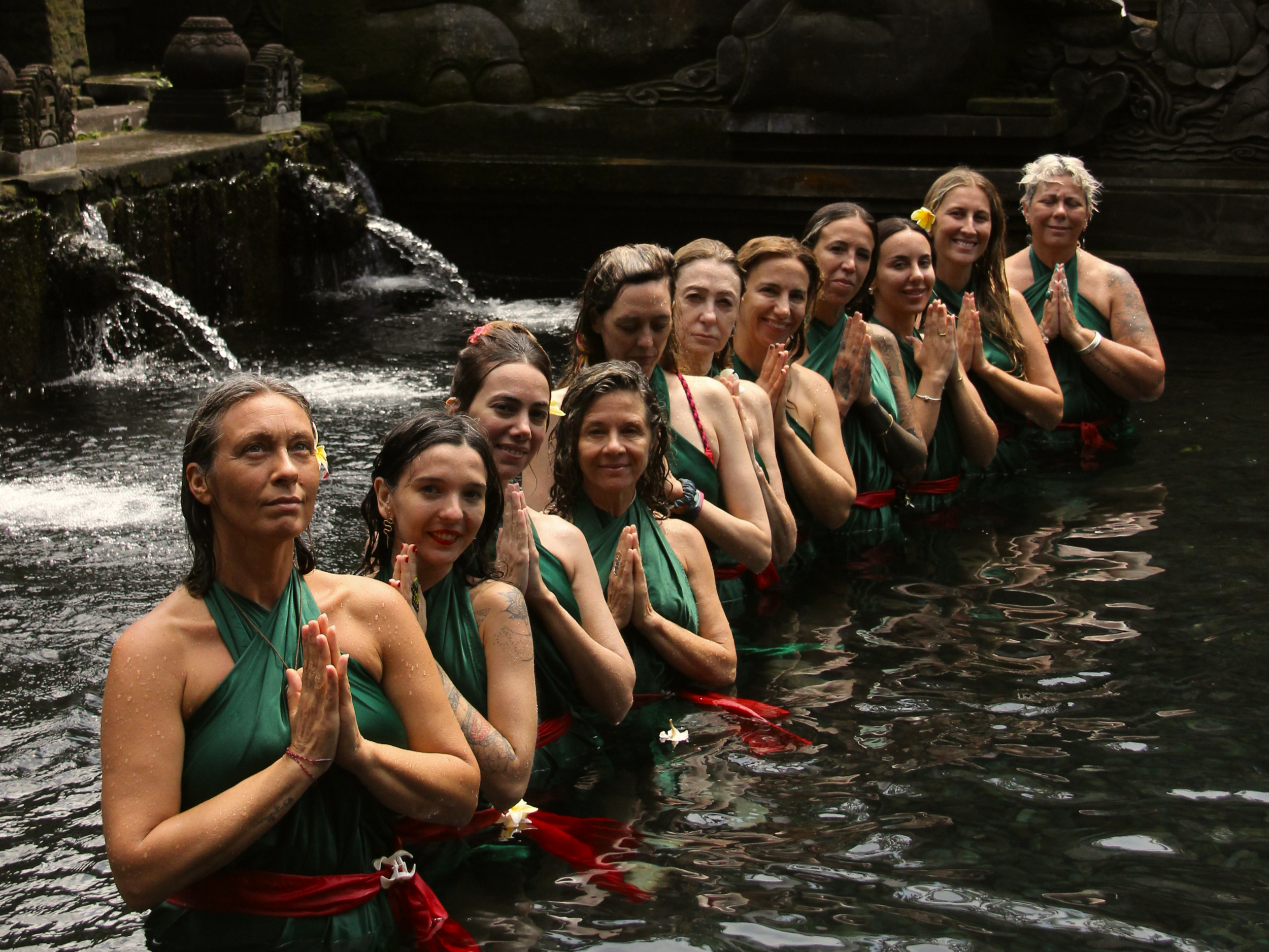 Grupo de mujeres participando en una ceremonia espiritual en un río oscuro, usando prendas verdes con cinturones rojos y flores en el cabello, en un entorno natural con chorros de agua y esculturas de piedra.