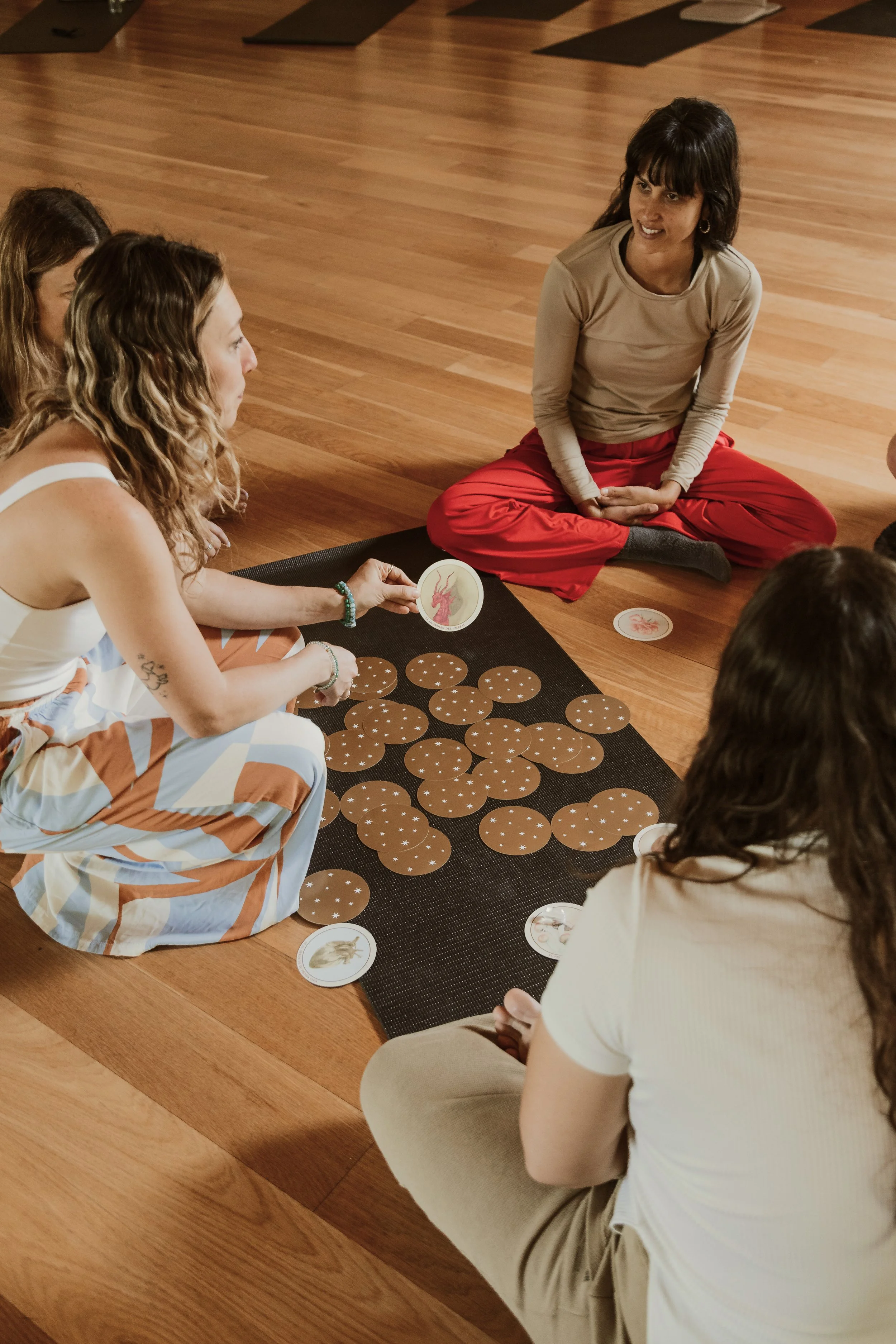 Un grupo de personas participando en un juego de mesa en un ambiente interior con suelo de madera, con una mujer sentada en el piso atendiendo a los demás.