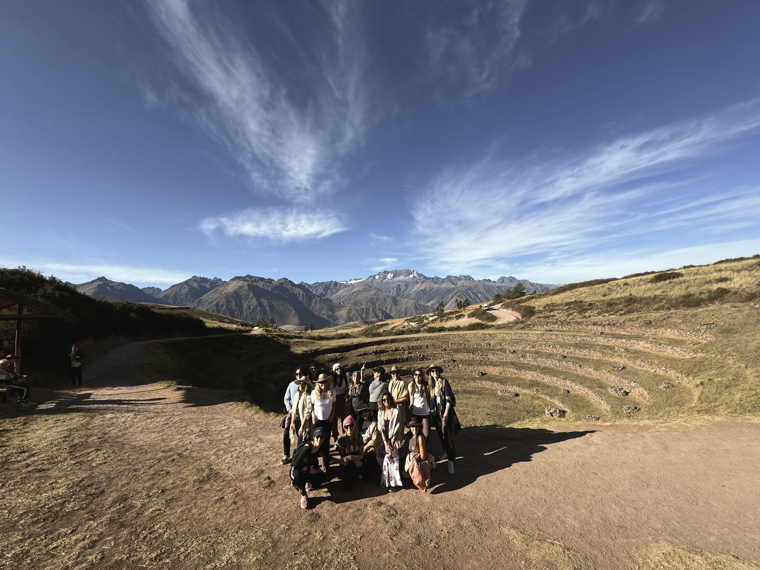 Grupo de personas en un sitio arqueológico en una zona montañosa con terrazas, con un cielo despejado y montañas al fondo.
