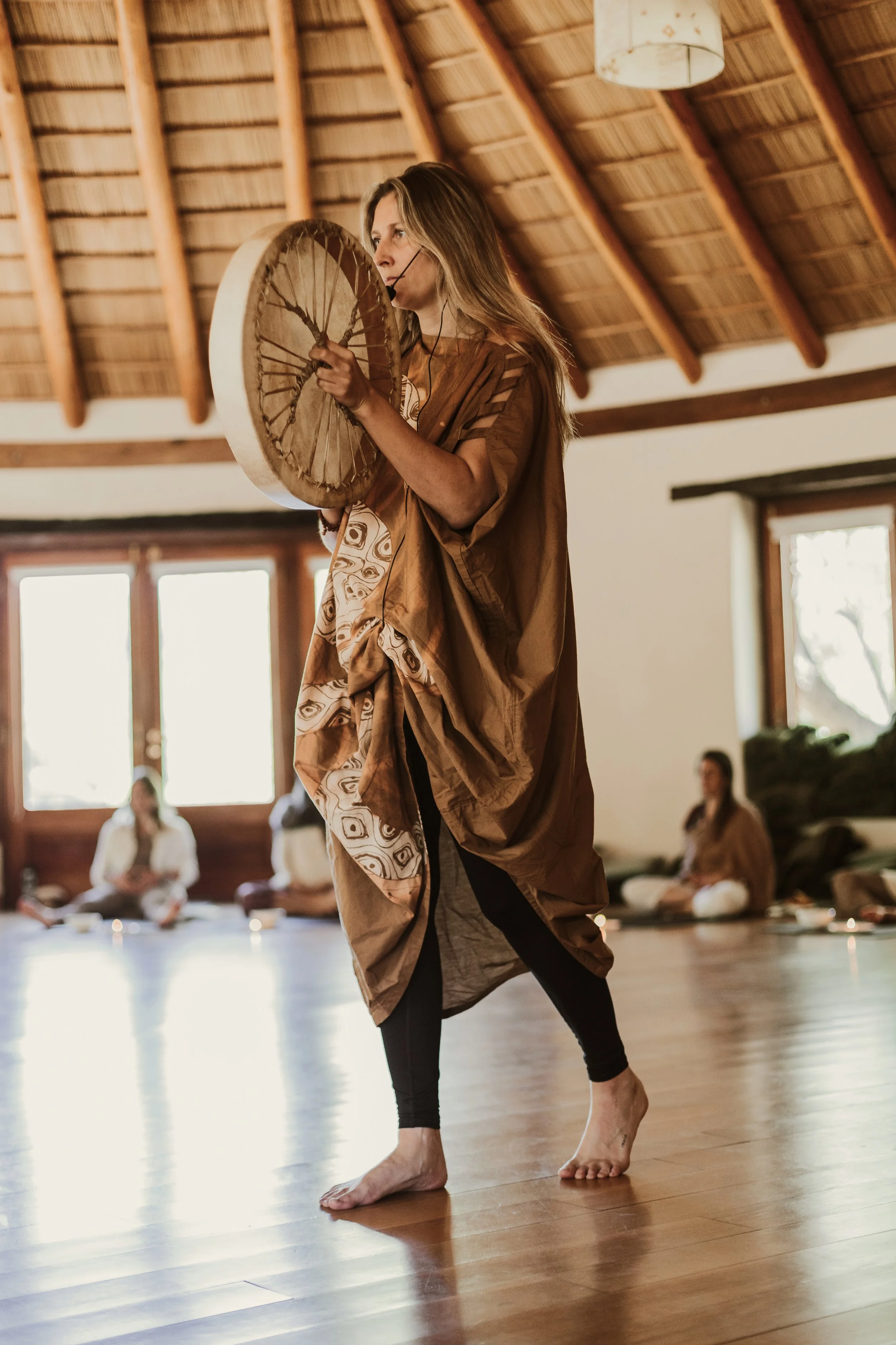 Mujer realizando danza con un tambor ceremonial en un entorno de interior con estructura de madera y varias personas sentadas en el fondo.
