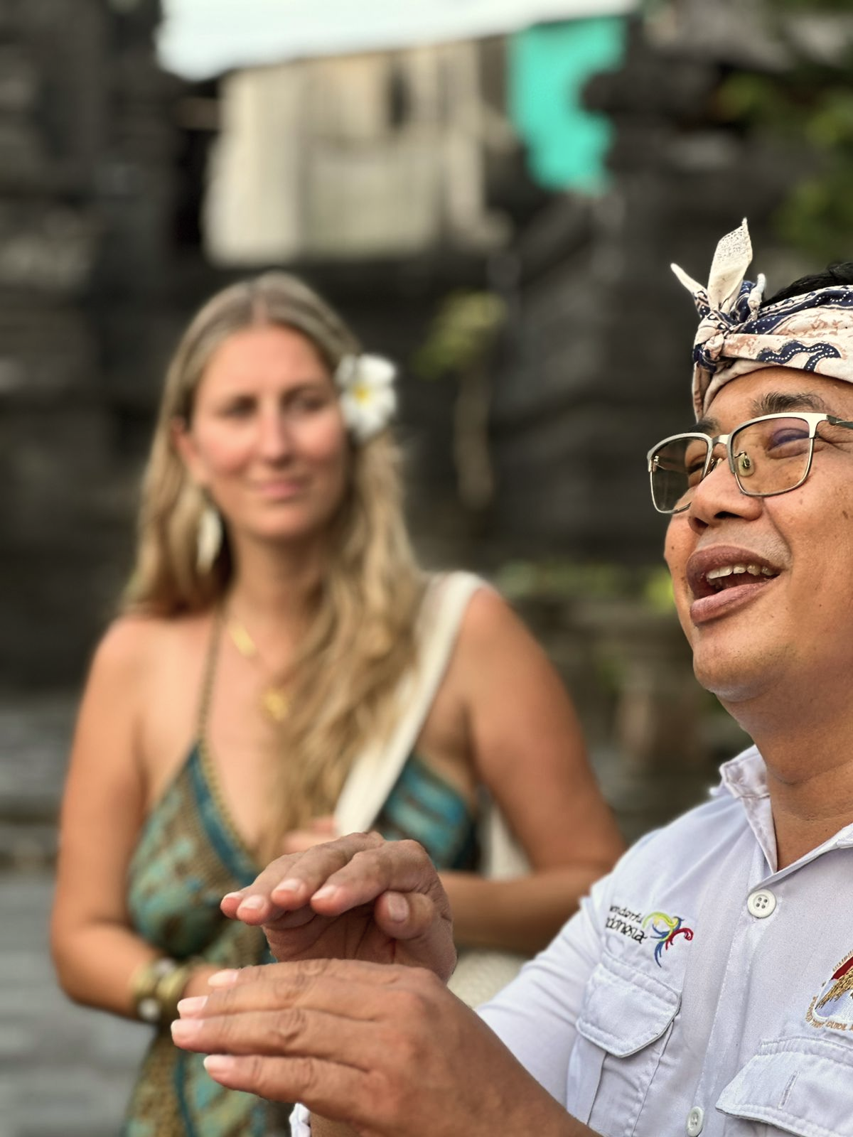 Hombre con gafas, bandana y camisa blanca conversando, mujer con vestido colorido y flor en el cabello en un entorno natural de piedra y vegetación.