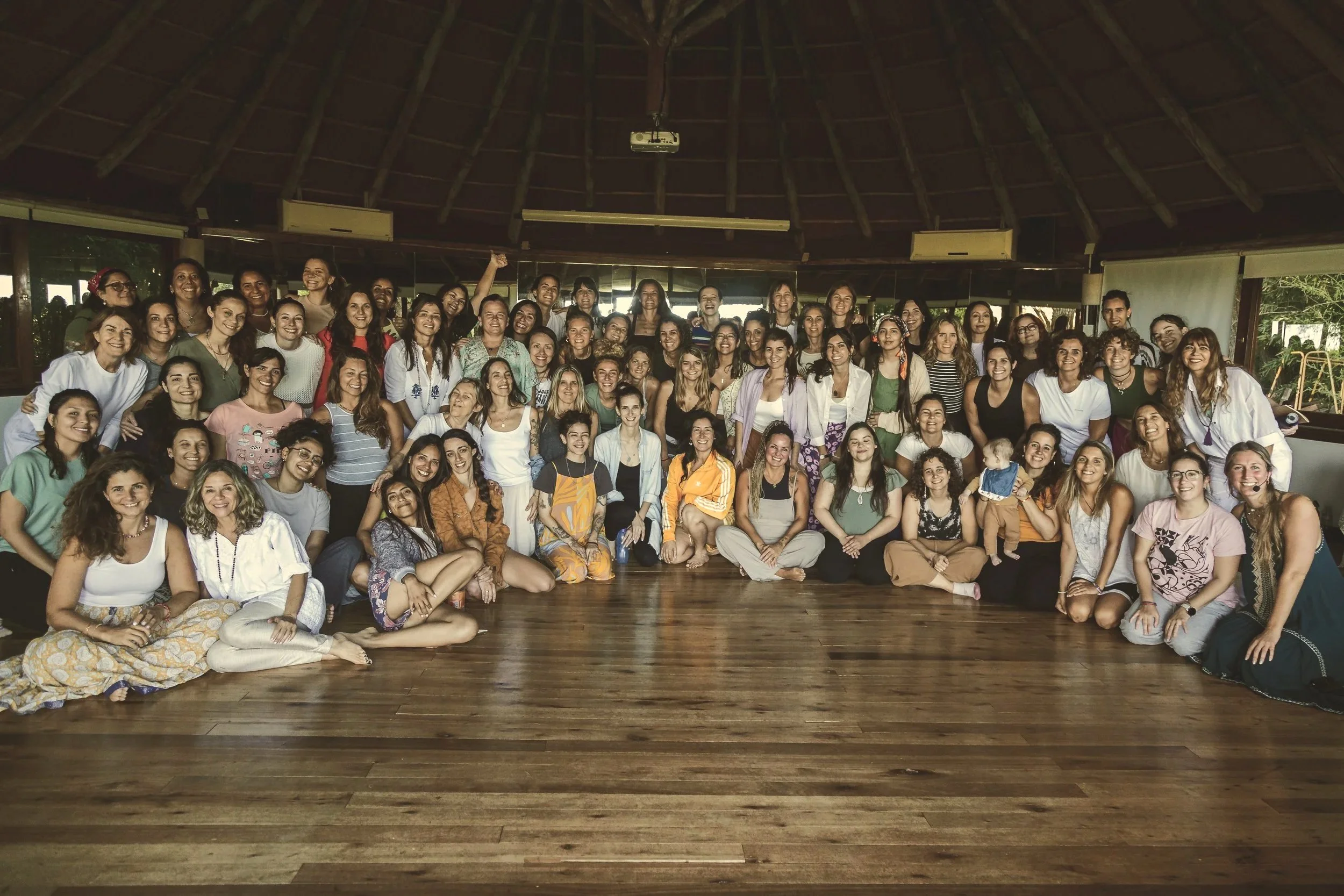Grupo grande de mujeres en una sala de madera con techo de palapa, algunas sonriendo y otras haciendo gestos, en un ambiente de convivencia amistosa.