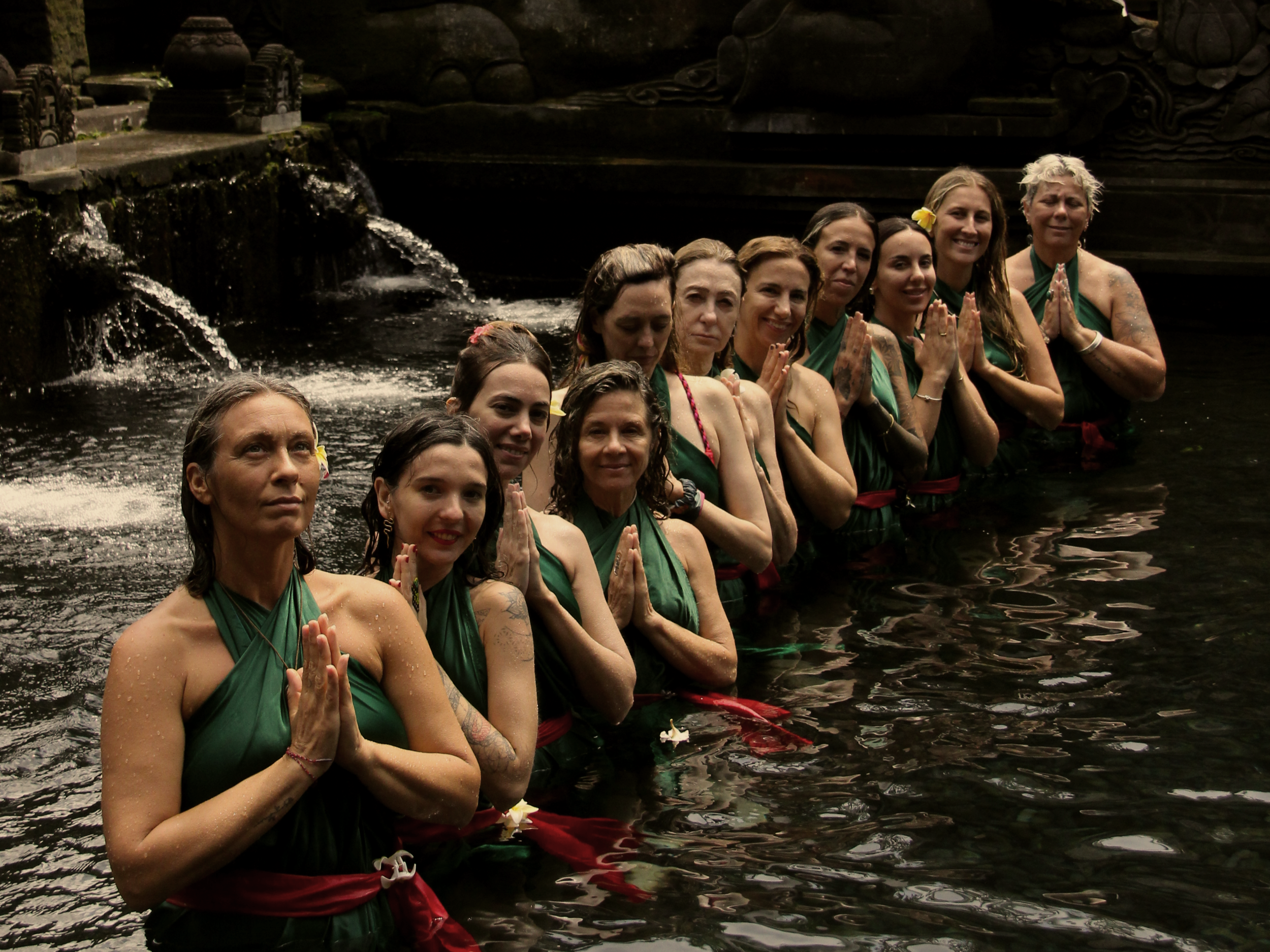 Grupo de mujeres en el agua en un ritual religioso en un templo, vestidas con atuendos verdes y fajas rojas, en una ceremonia espiritual.