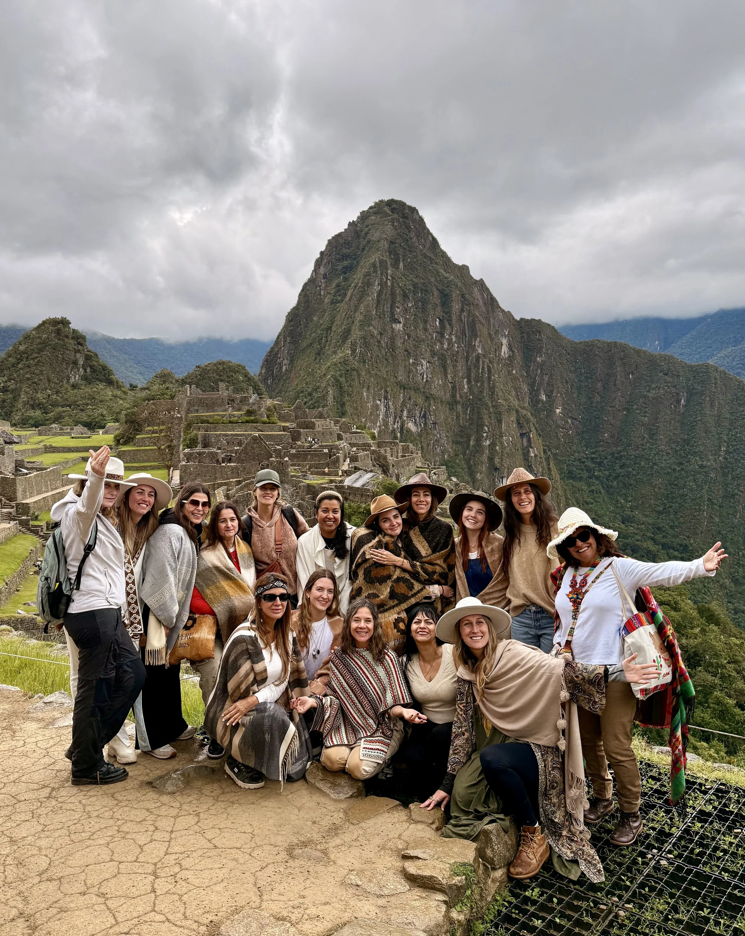 Grupo de turistas en Machu Picchu, rodeados de montañas y ruinas antiguas, con vestimenta de viaje y sombreros, en un día nublado.