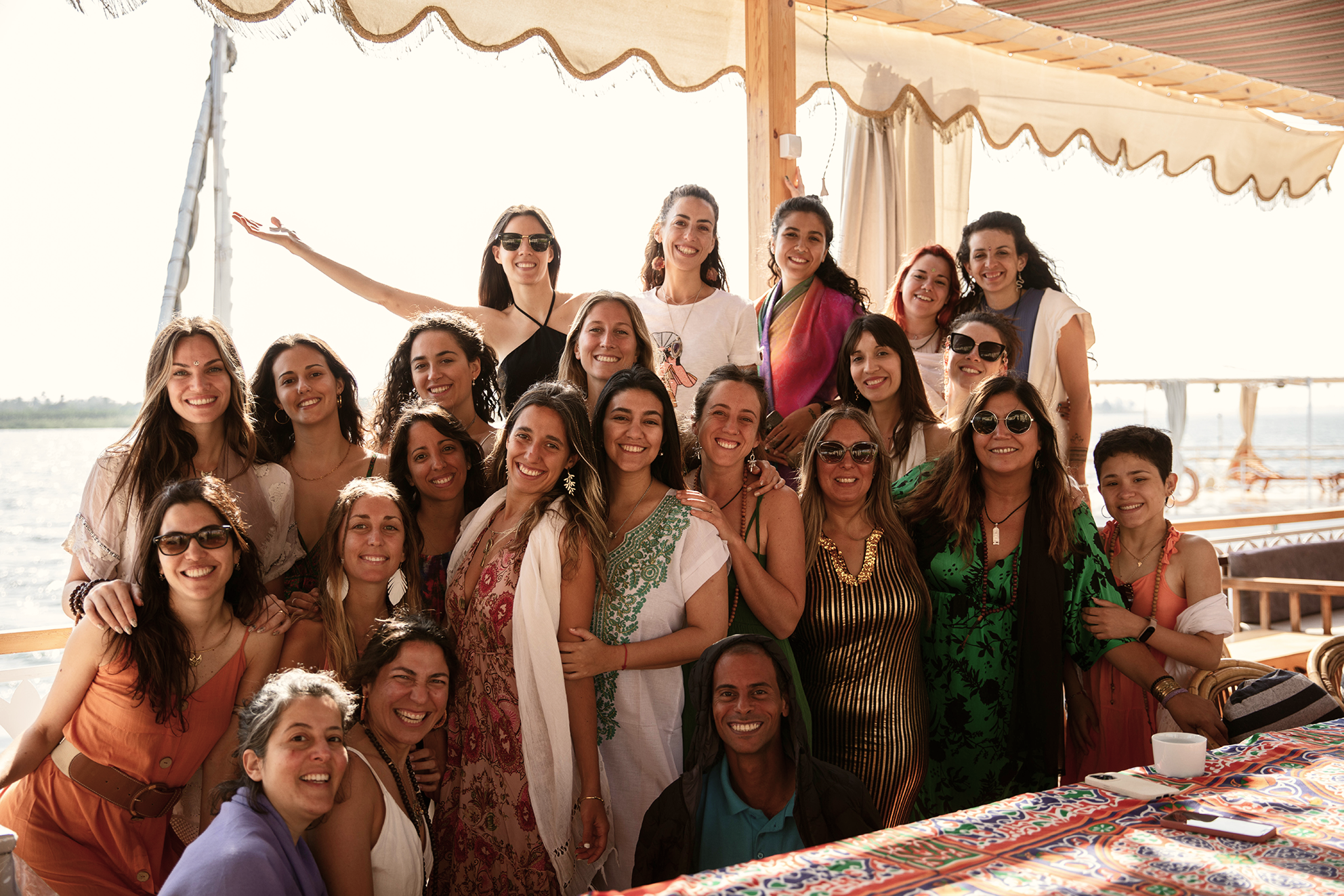 Grupo de mujeres y un niño posando en una terraza junto a un río, durante el día, con decoración de toldo y mesas, en un ambiente alegre y soleado.