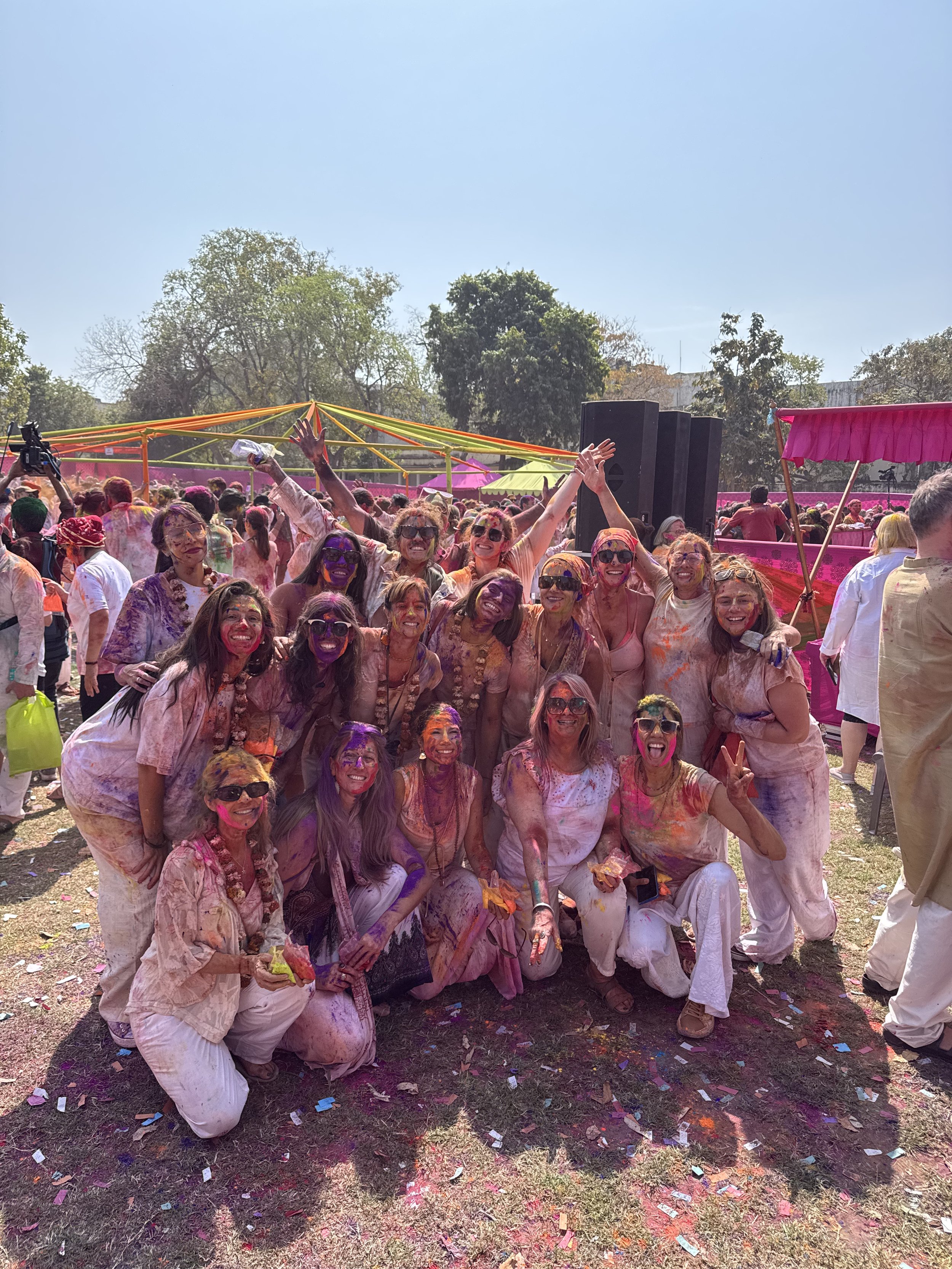 Grupo de personas celebrando en un festival de colors, cubiertas de polvo de colores, sonriendo y levantando las manos, con un fondo de árboles y un cielo azul.