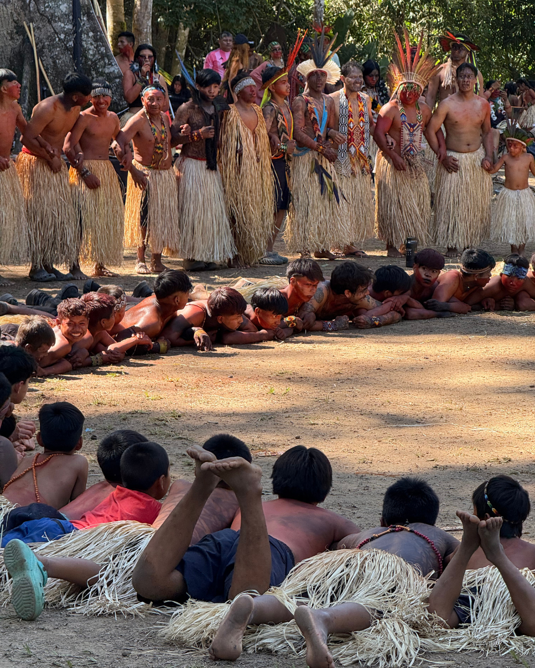 Grupo de personas indígenas en vestimenta tradicional, participando en un baile o ceremonia en la naturaleza, con varias personas acostadas en el suelo observando.