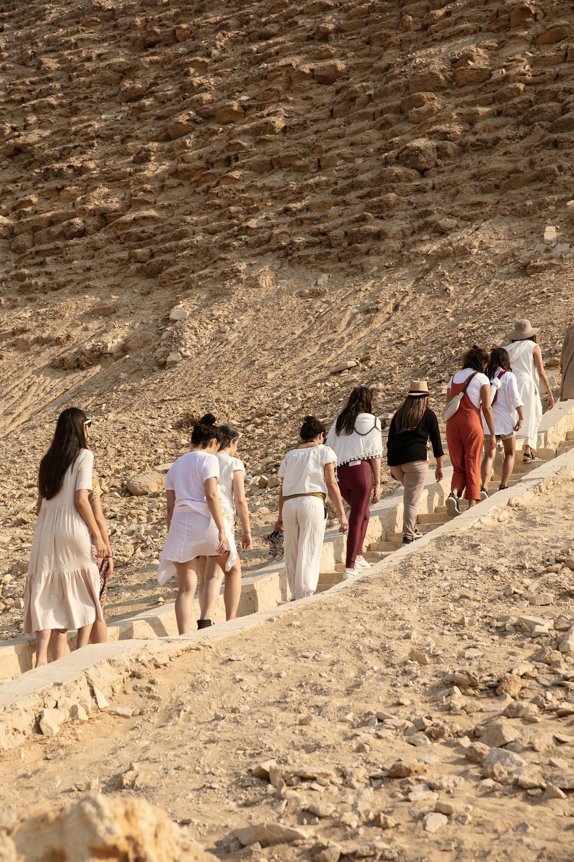 Grupo de personas caminando en una rampa en un entorno desértico con rocas y tierra, algunos con sombrero y ropa ligera.