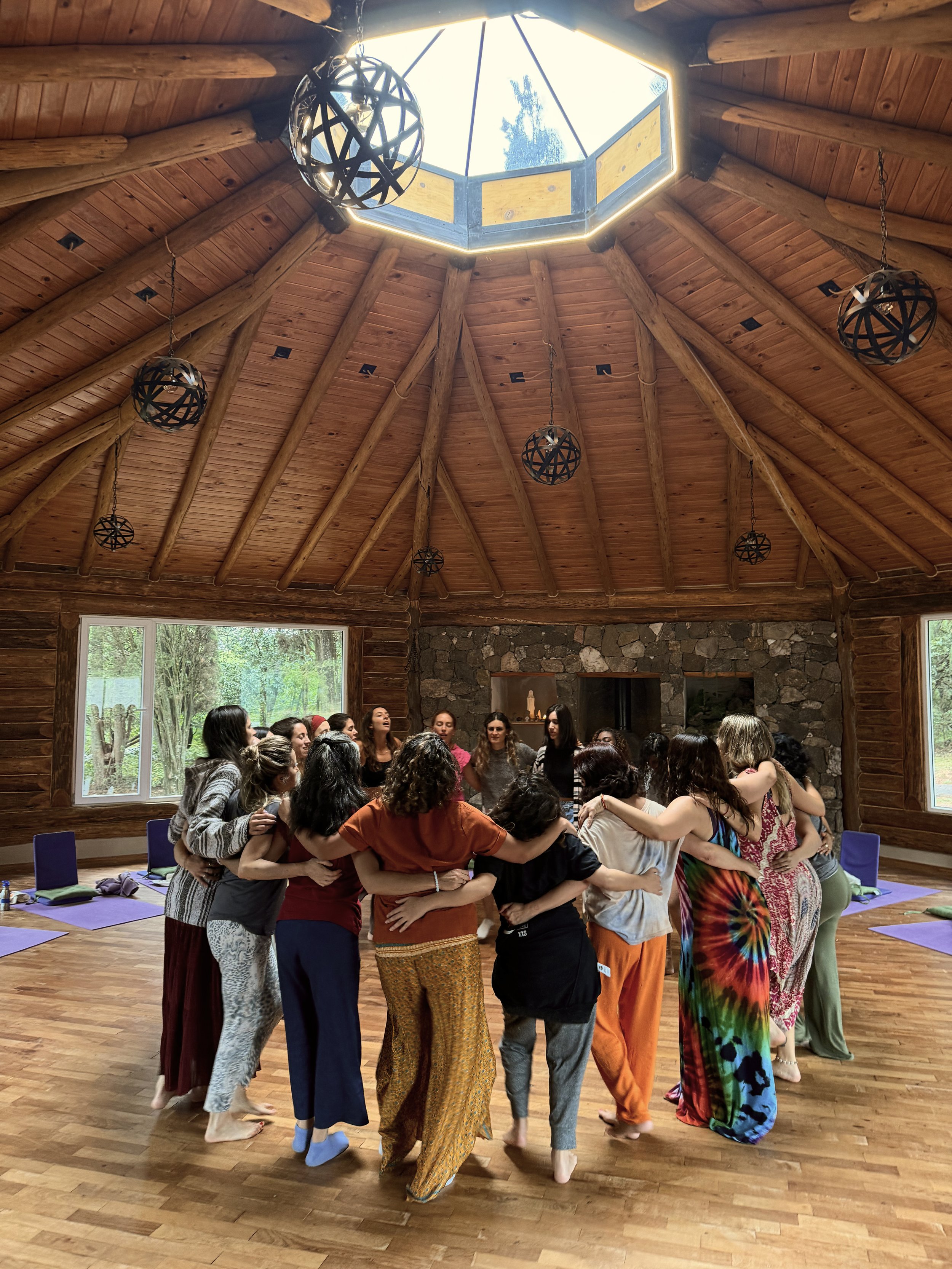 Grupo de personas en un círculo, abrazándose, en una sala de madera con ventanas y techos en forma de cúpula, con luces colgantes y alfombra de yoga en el suelo.
