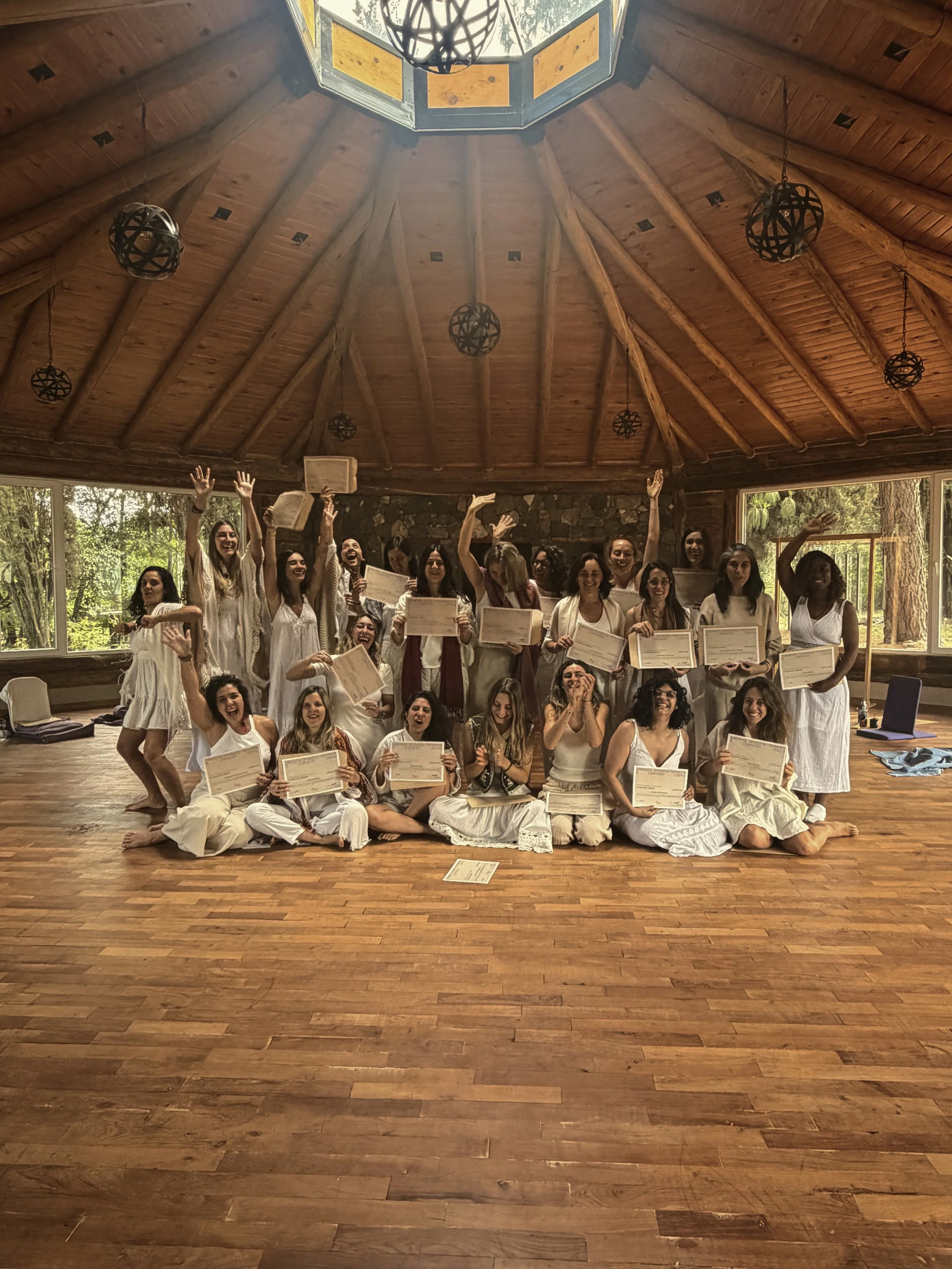 Grupo de mujeres en una ceremonia de graduación en una sala de madera, todas sosteniendo certificados y luciendo felices.