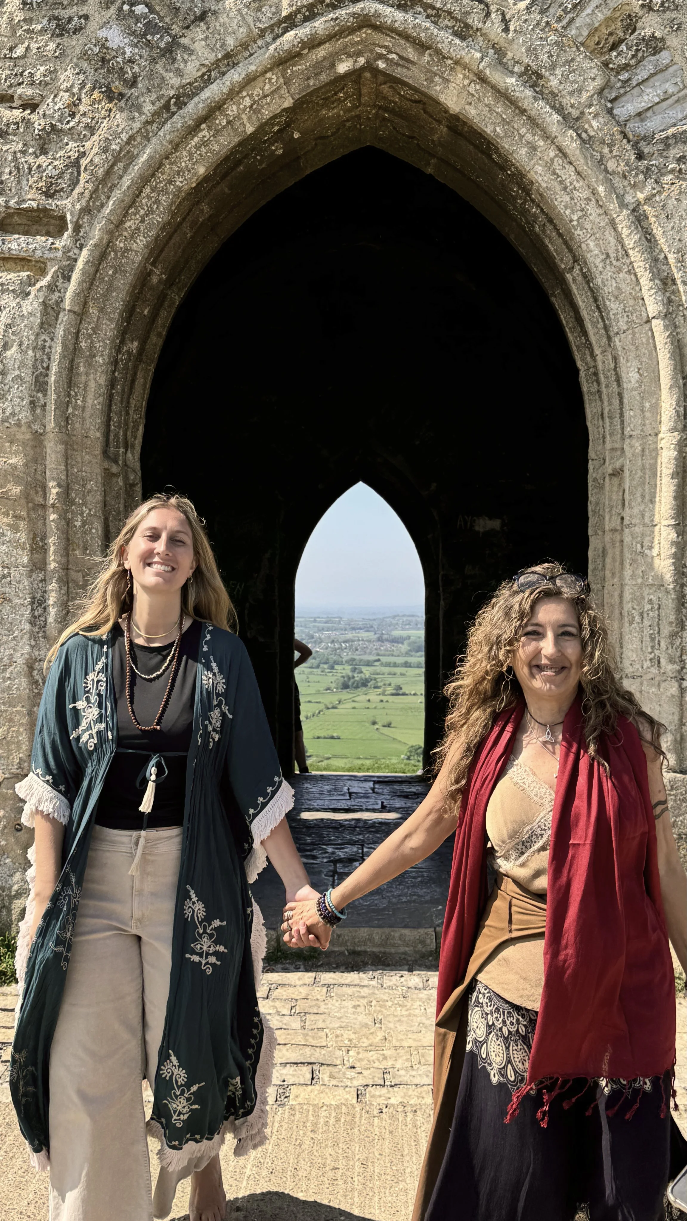 Dos mujeres de espaldas, tomadas de la mano, posando en un marco de ventana en ruinas con vista al campo y al cielo despejado.