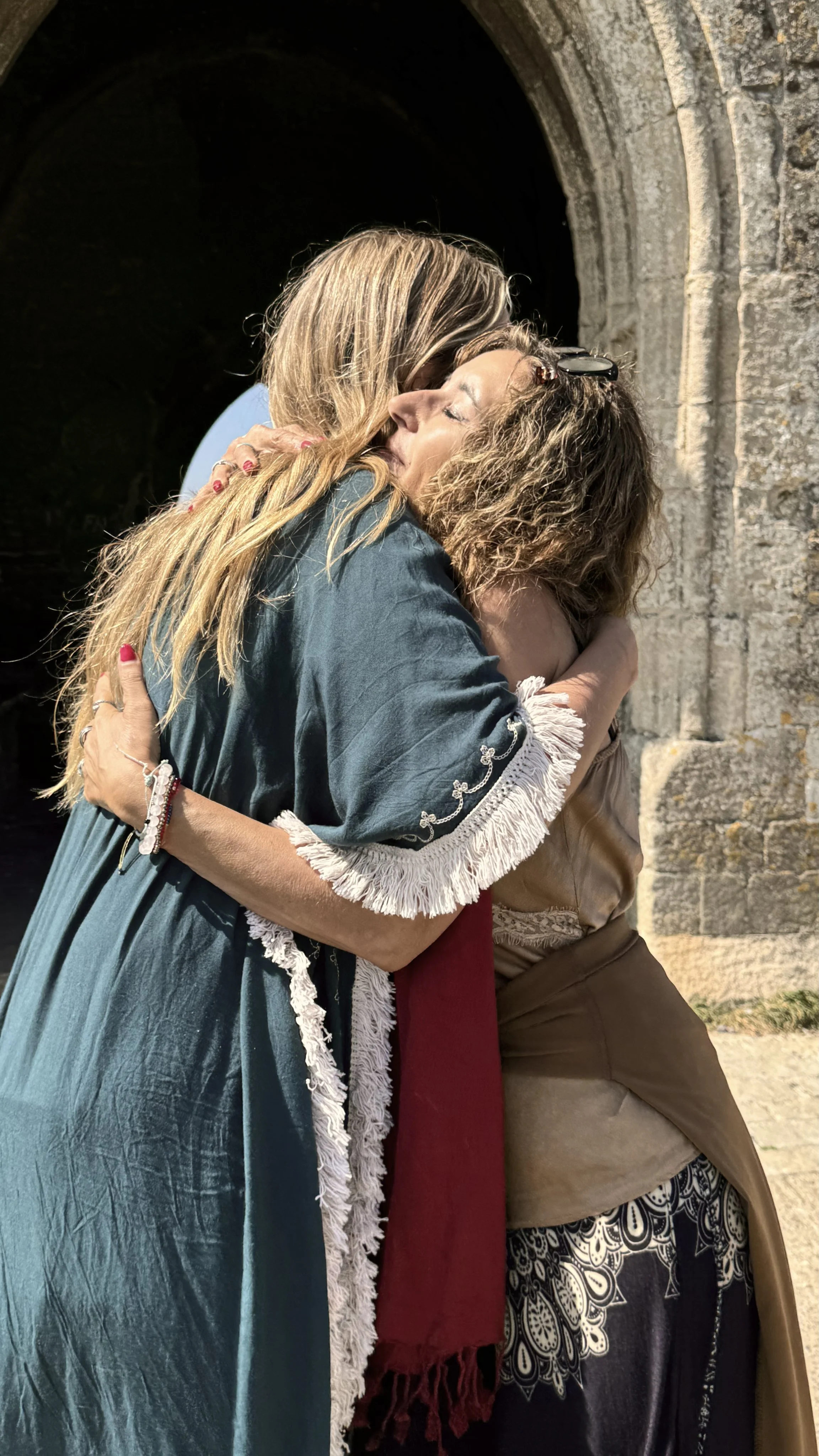 Dos mujeres dándose un abrazo frente a un arco de piedra, una con cabello rubio y otra con cabello rizado, en un entorno exterior.