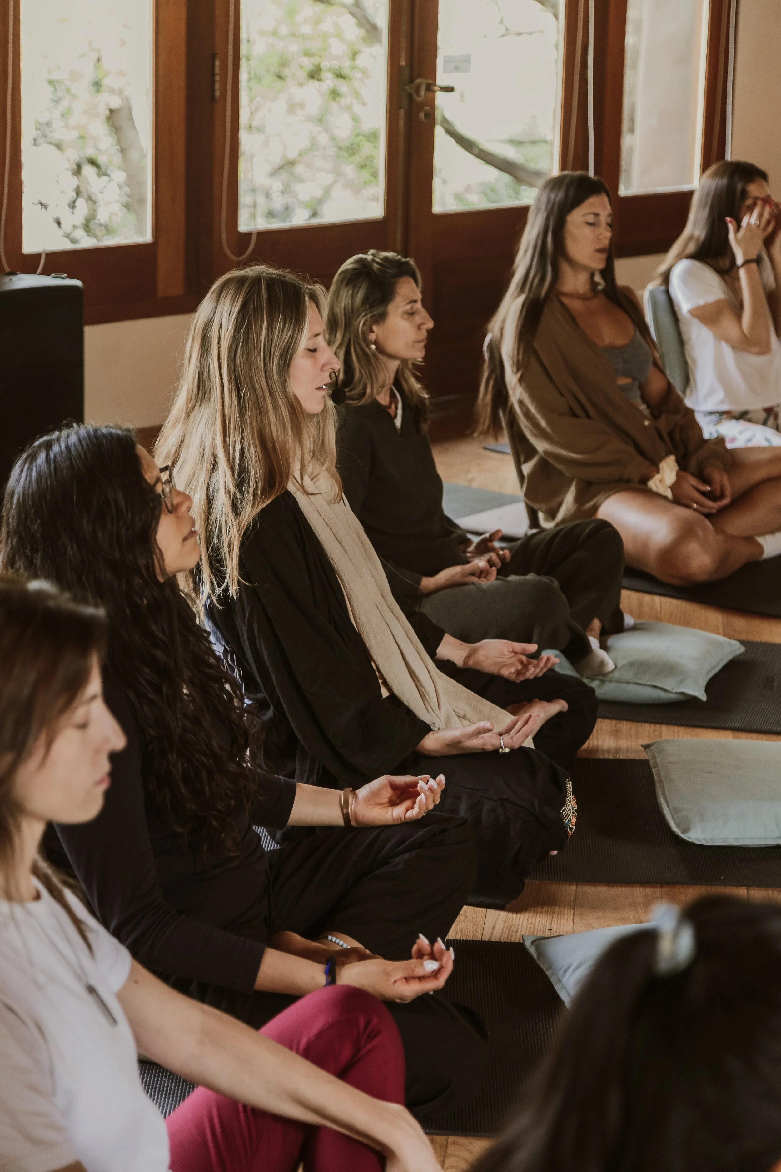 Grupo de mujeres en posición de meditación en una habitación con ventanas de madera y vista a árboles.