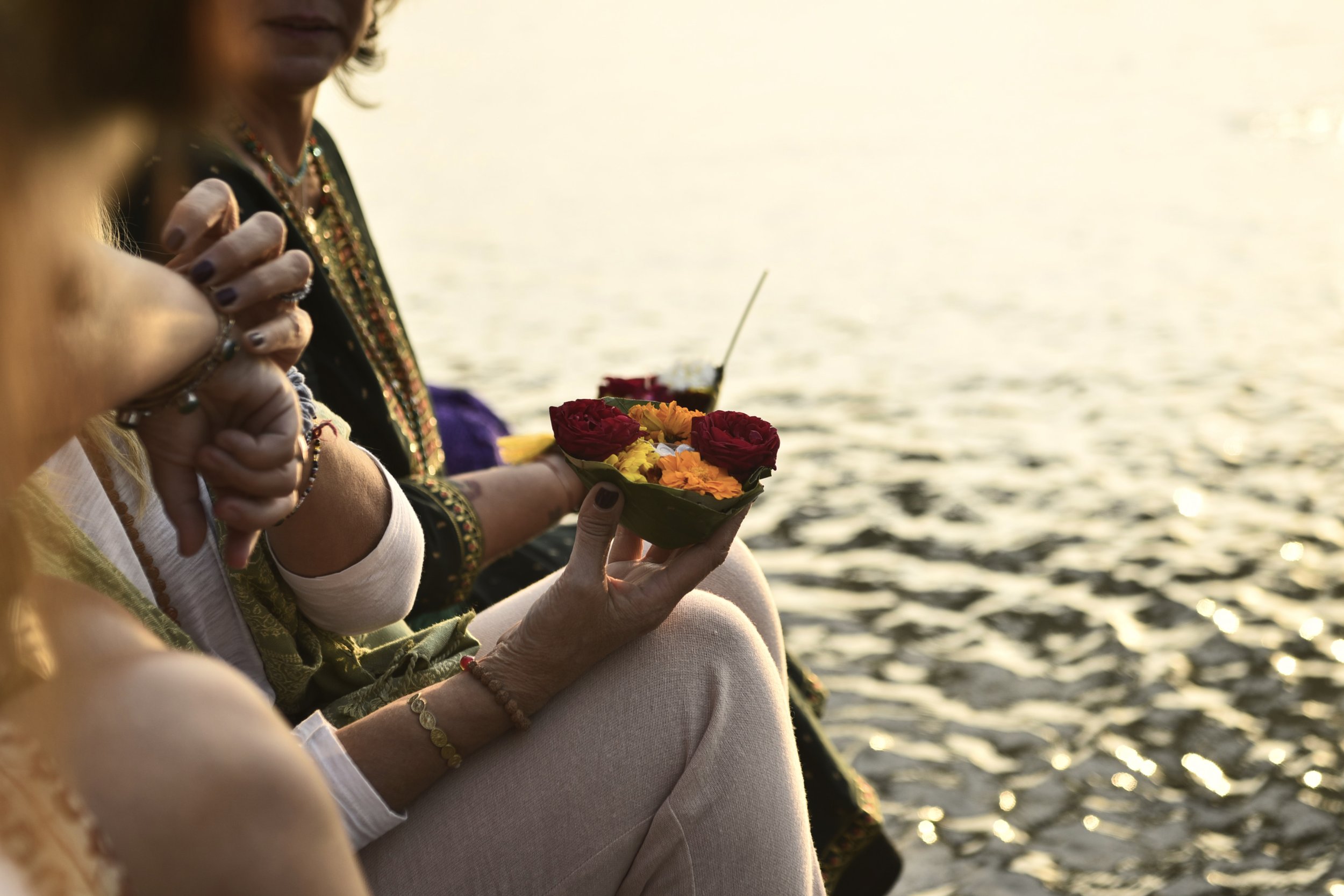 Personas en un entorno al atardecer, una de ellas sosteniendo un plato con flores coloridas junto a un cuerpo de agua.
