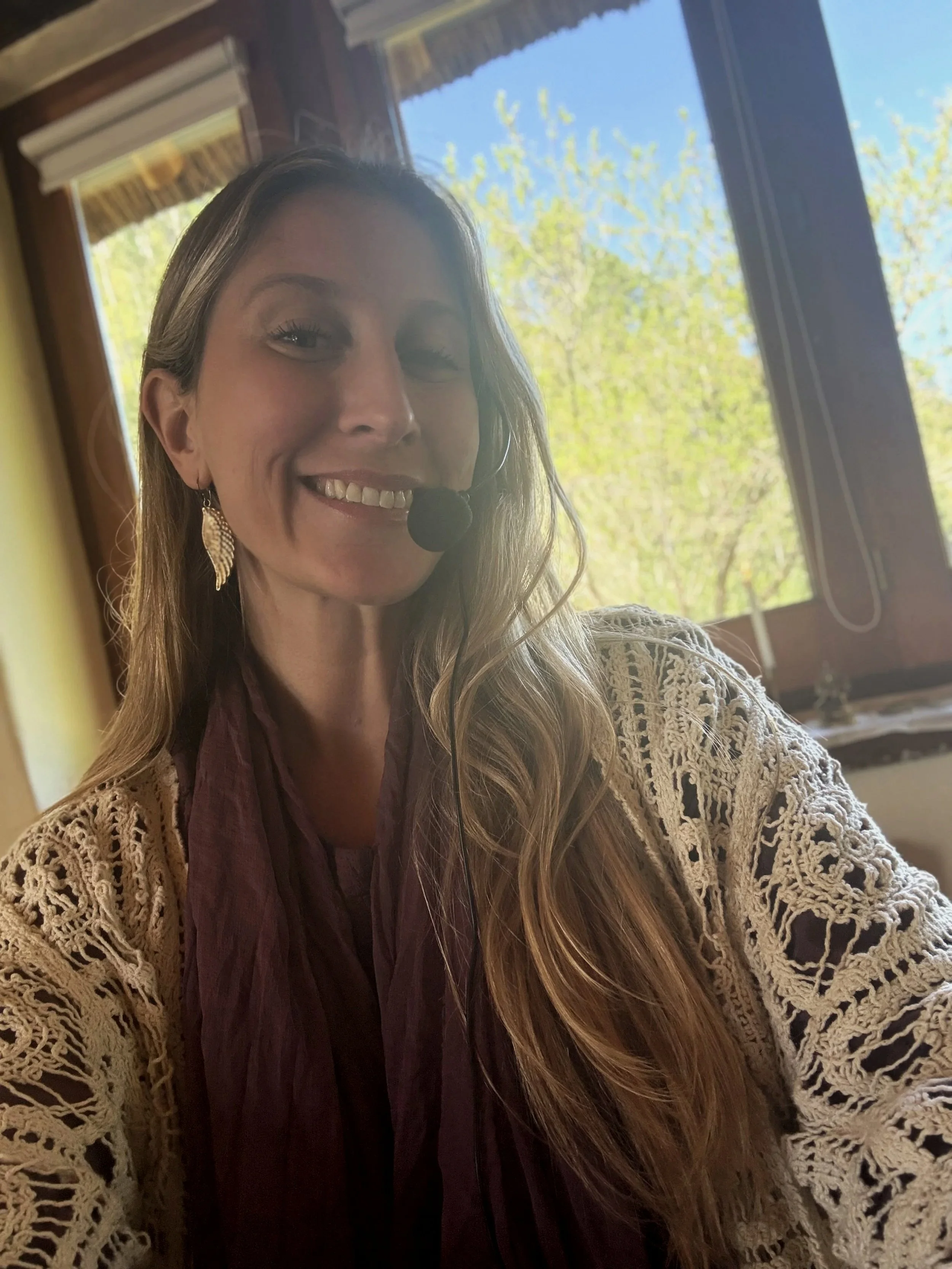 Mujer sonriendo con auriculares con micrófono, cabello largo y ondulado, ropa clara con un chaleco de ganchillo, en un interior con ventanas y vista a árboles verdes en un día soleado.