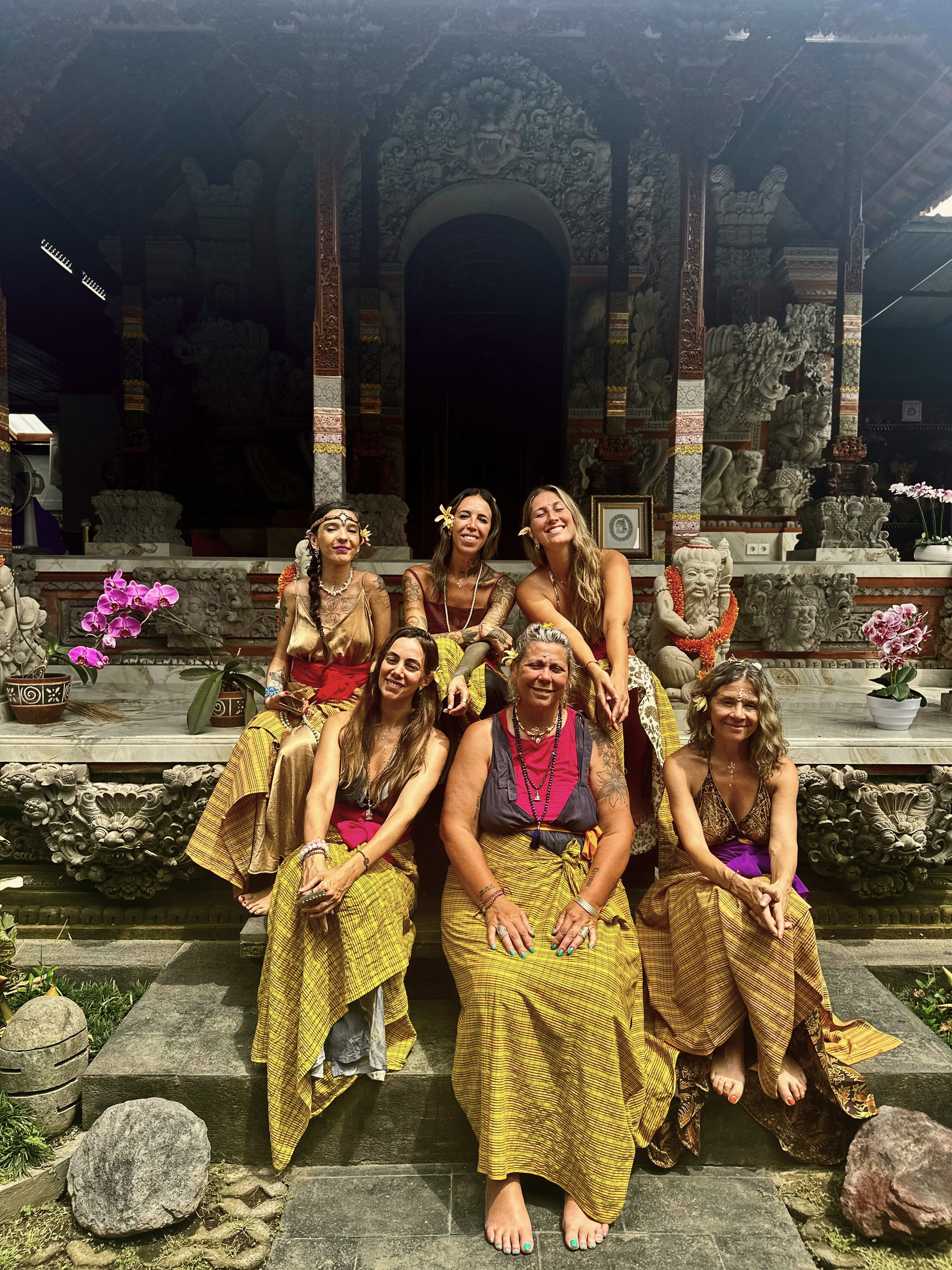 Grupo de siete mujeres sentadas y posando en un santuario con esculturas de piedra, flores y detalles culturales, vestidas con ropa tradicional y accesorios en un ambiente artístico y religioso.