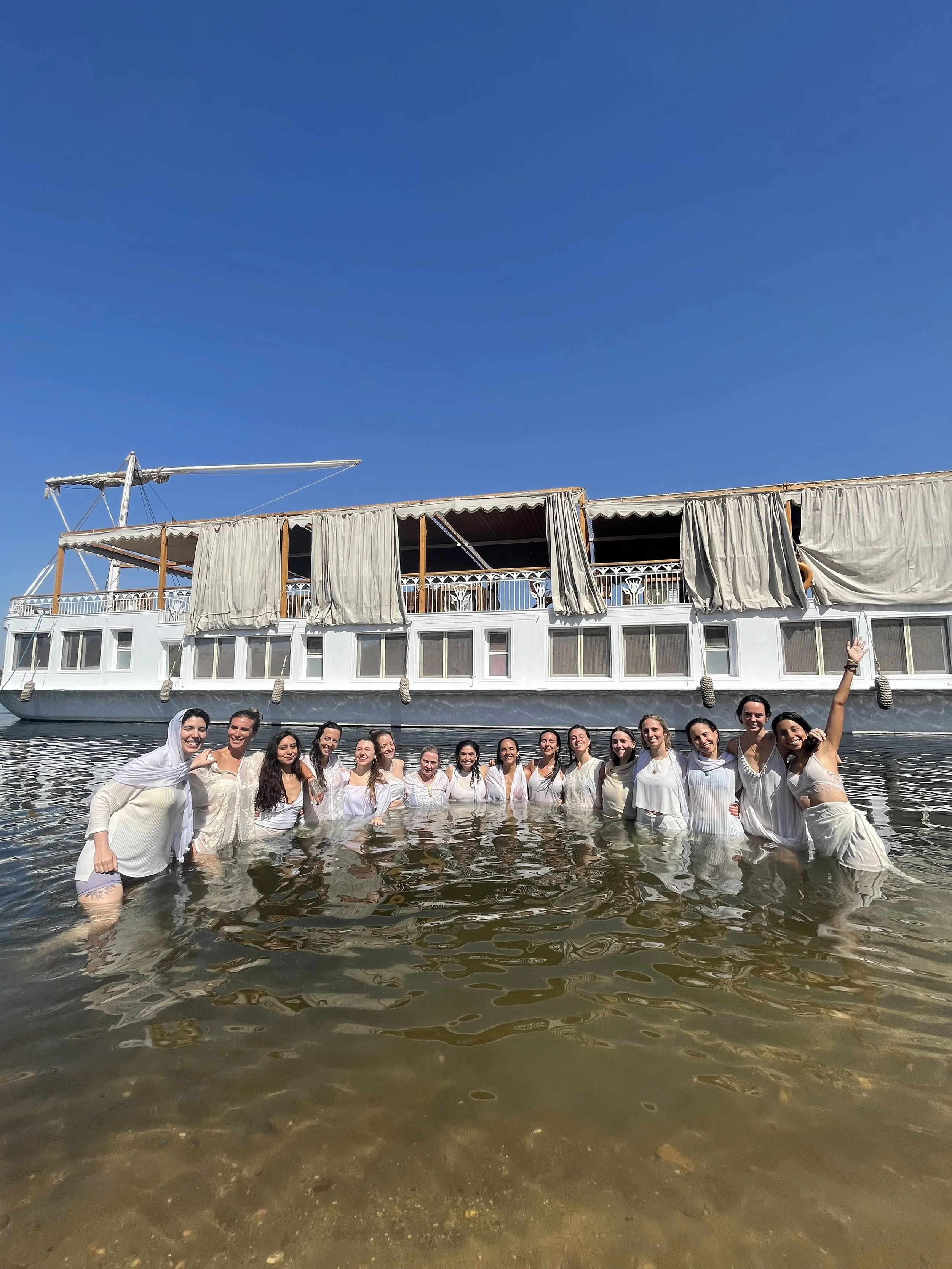 Un grupo de mujeres vestidas de blanco en el agua frente a un barco de madera en un día soleado y cielo despejado.
