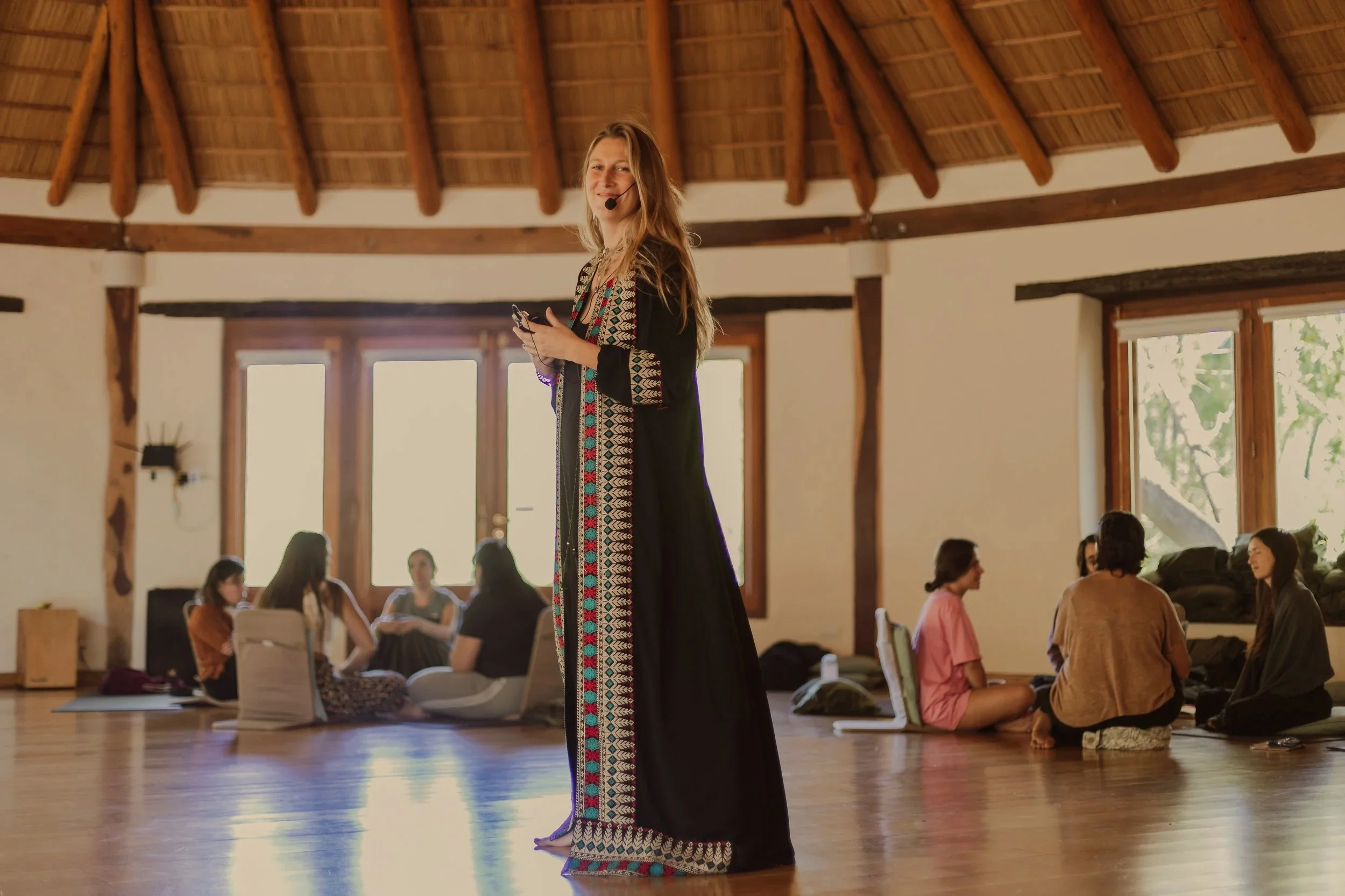 Mujer en un taller o clase de yoga o meditación en un espacio amplio con ventanas grandes y techo de madera, algunas personas sentadas en el suelo conversando o participando en la actividad.