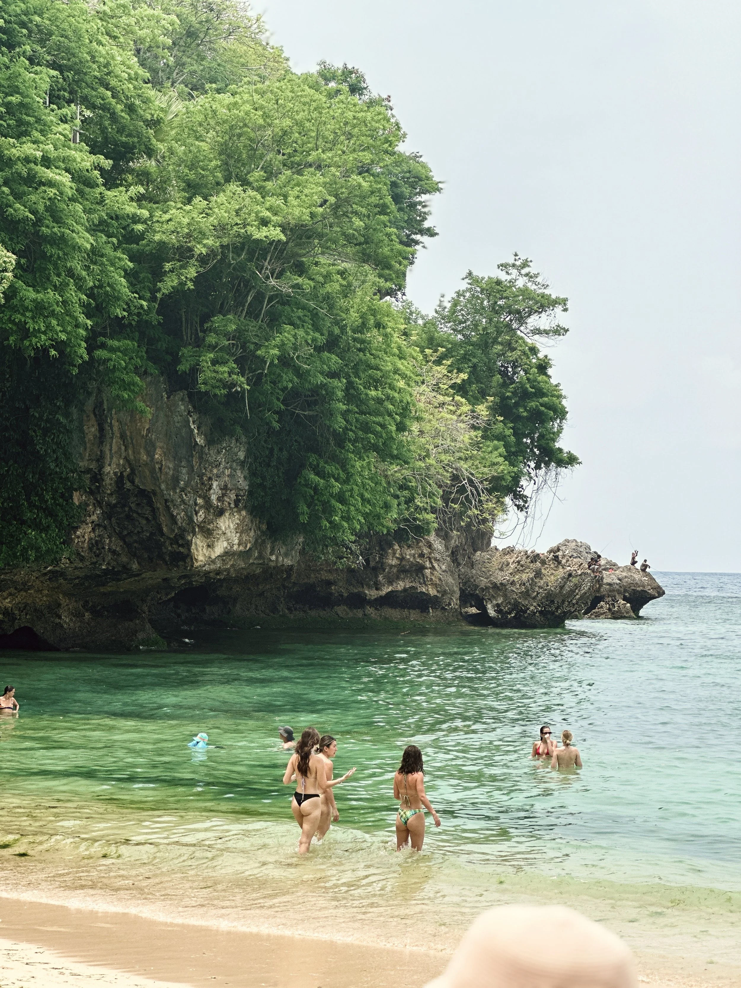 Personas en la playa con rocas y vegetación exuberante en un acantilado.