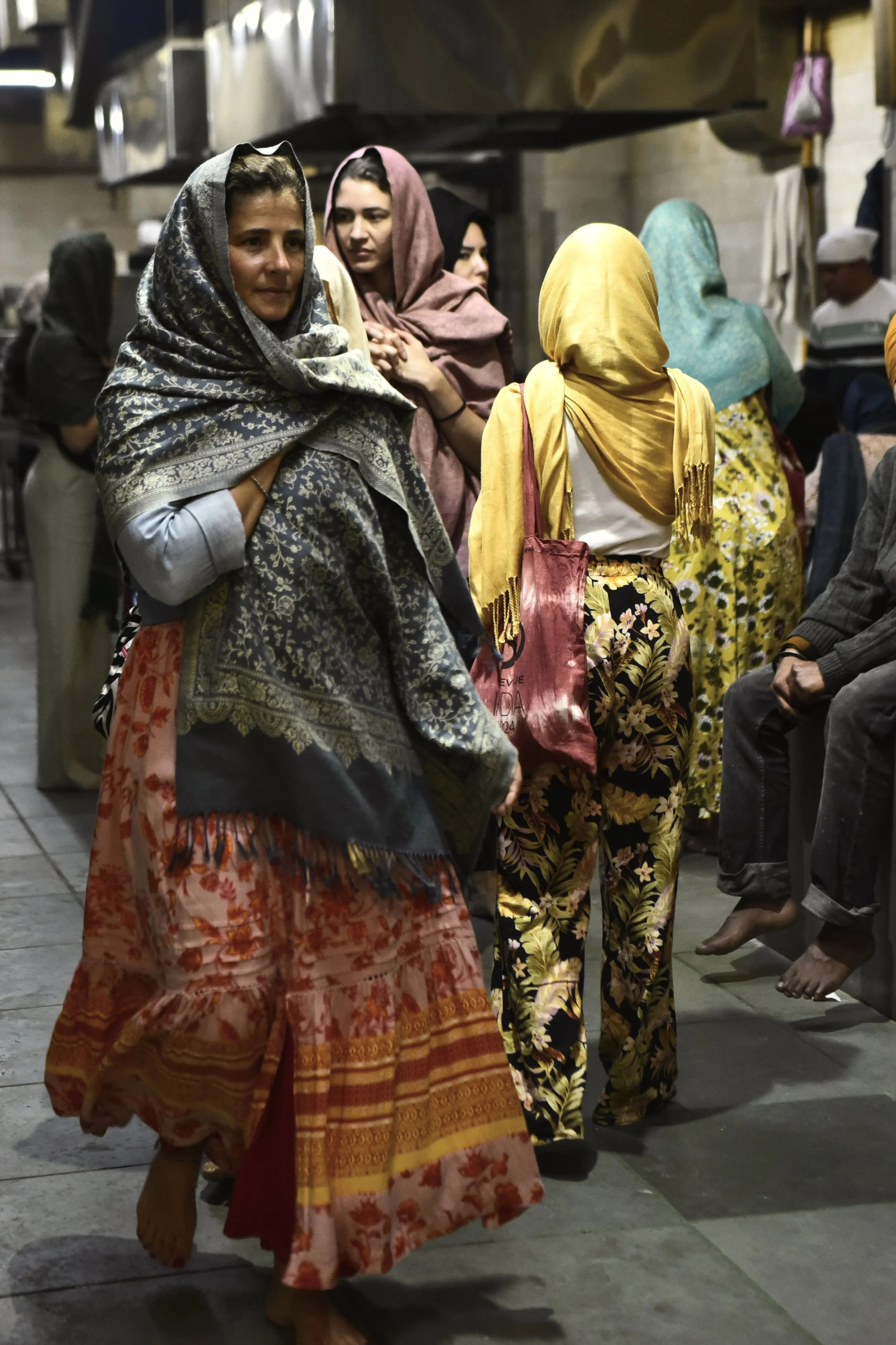 Grupo de mujeres con pañuelos en la cabeza en una cocina comunitaria, algunas sentadas y otras de pie, con delantal y ropa colorida.