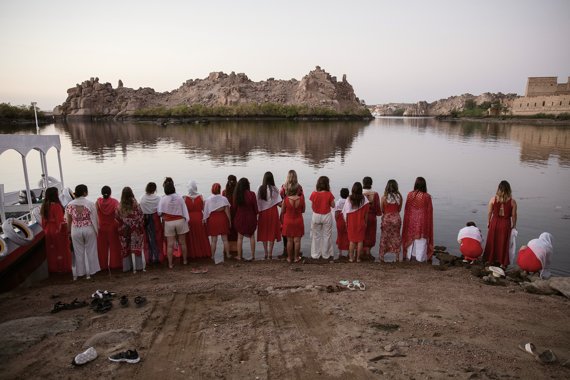 Grupo de personas de espaldas en la orilla de un río, vestidas mayormente con ropa roja y blanca, observando el agua durante el atardecer.