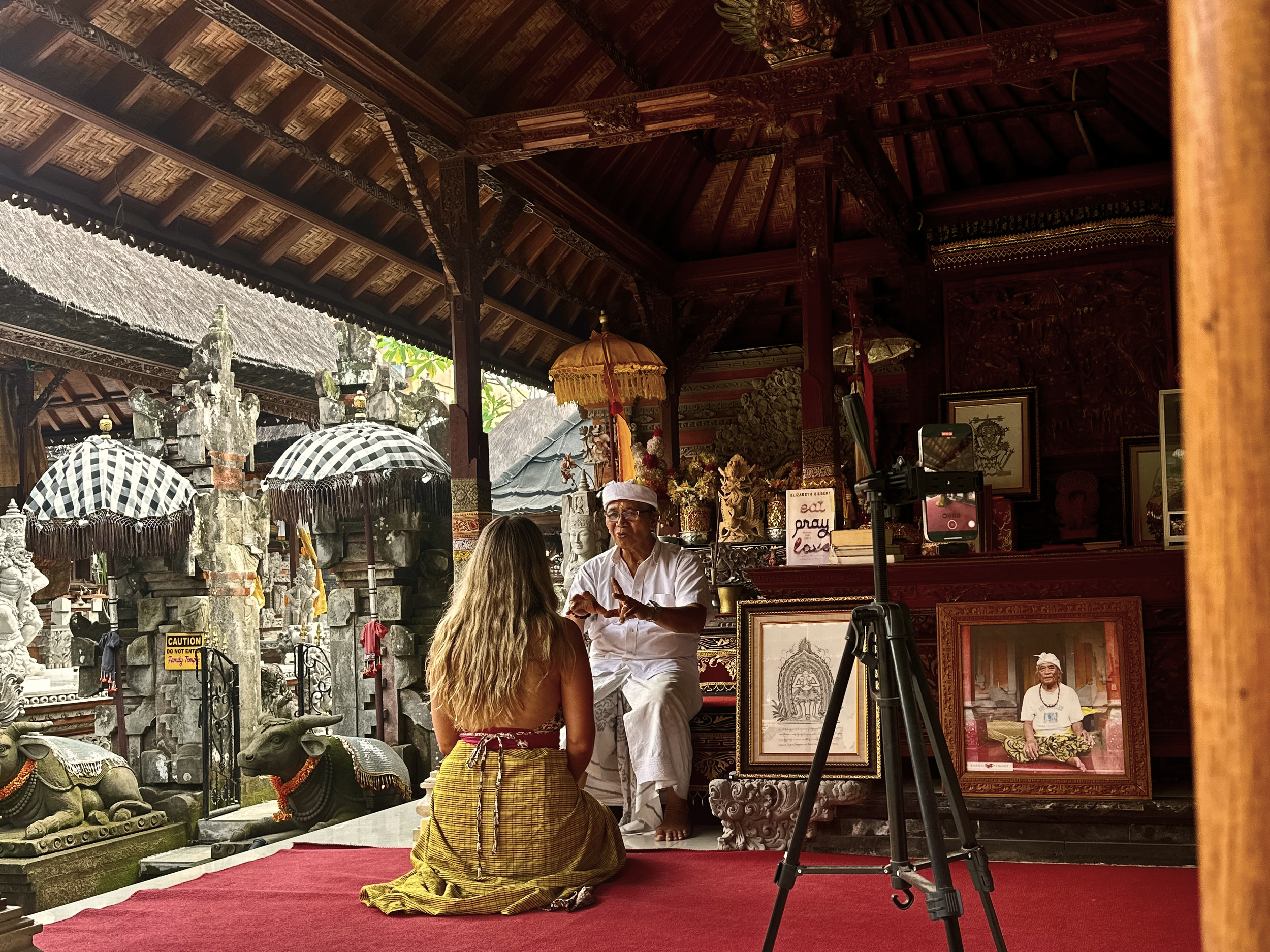 Un hombre vestido con ropa tradicional balinesa realiza un ritual en un templo, frente a una mujer que está de rodillas con ropa tradicional, en un espacio decorado con arte y fotos, en Bali, Indonesia.