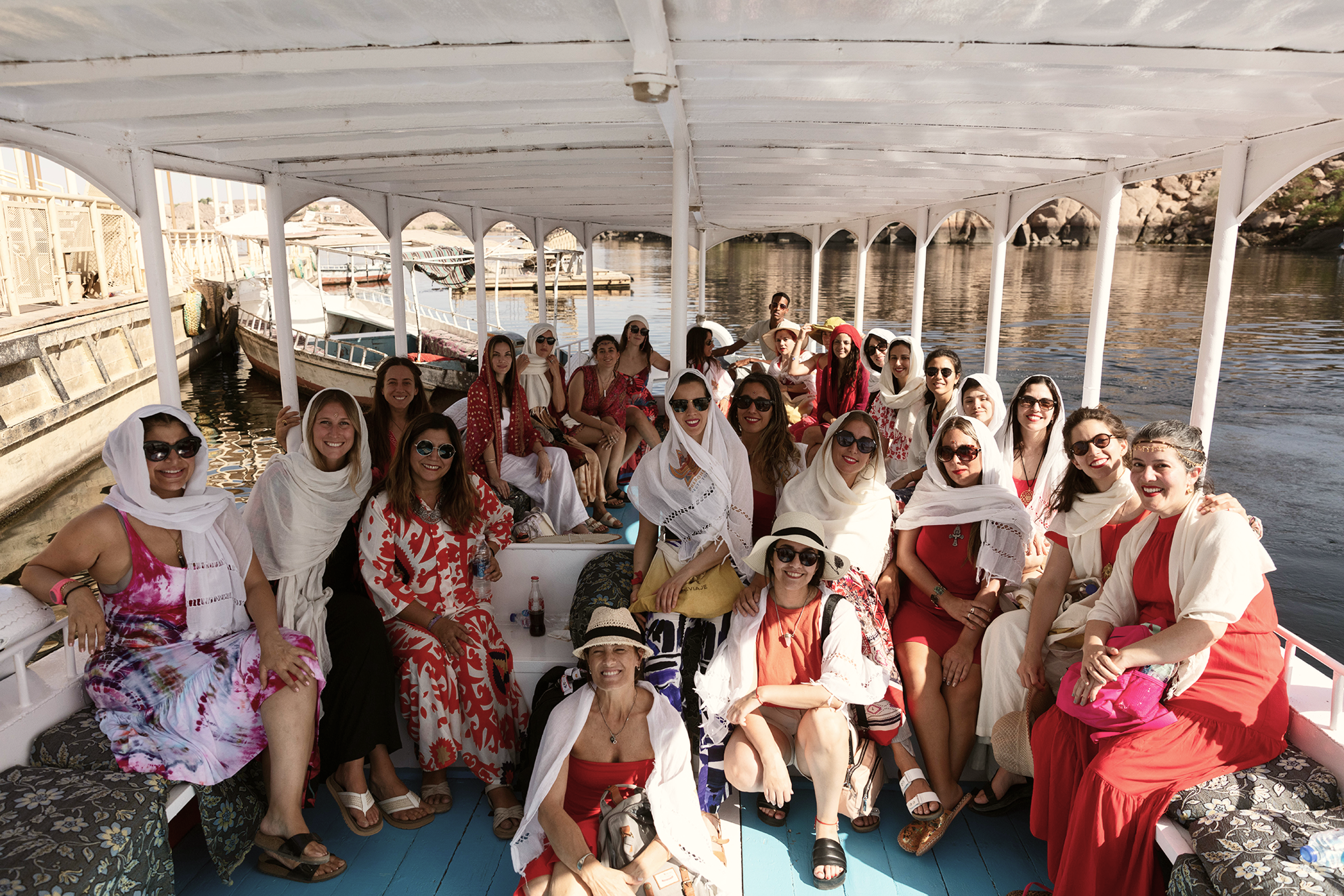 Grupo de mujeres turistas en un bote por un río, algunas llevan sombreros y gafas de sol, vestidas con ropa de verano en tonos rojos, blancos y colores claros, disfrutando de un tour en barco.