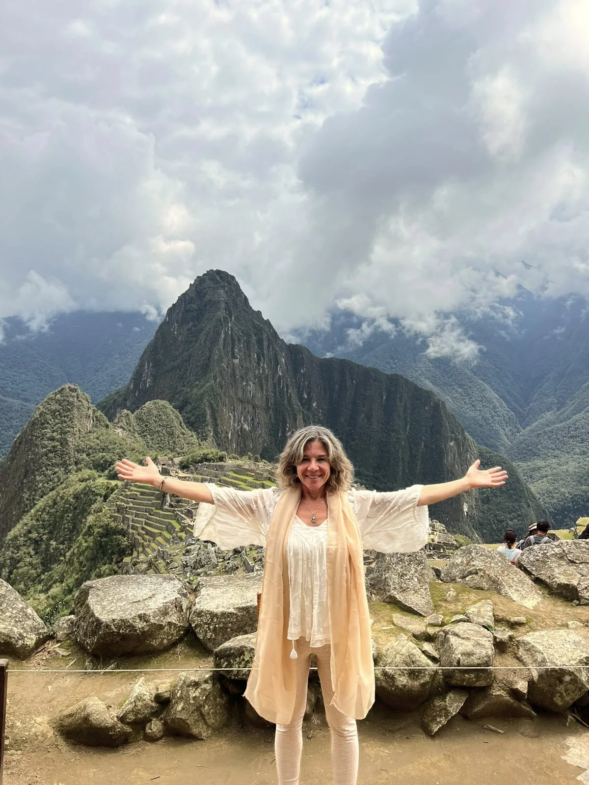 Mujer sonriendo con los brazos abiertos en Machu Picchu, con montañas y nubes en el fondo.