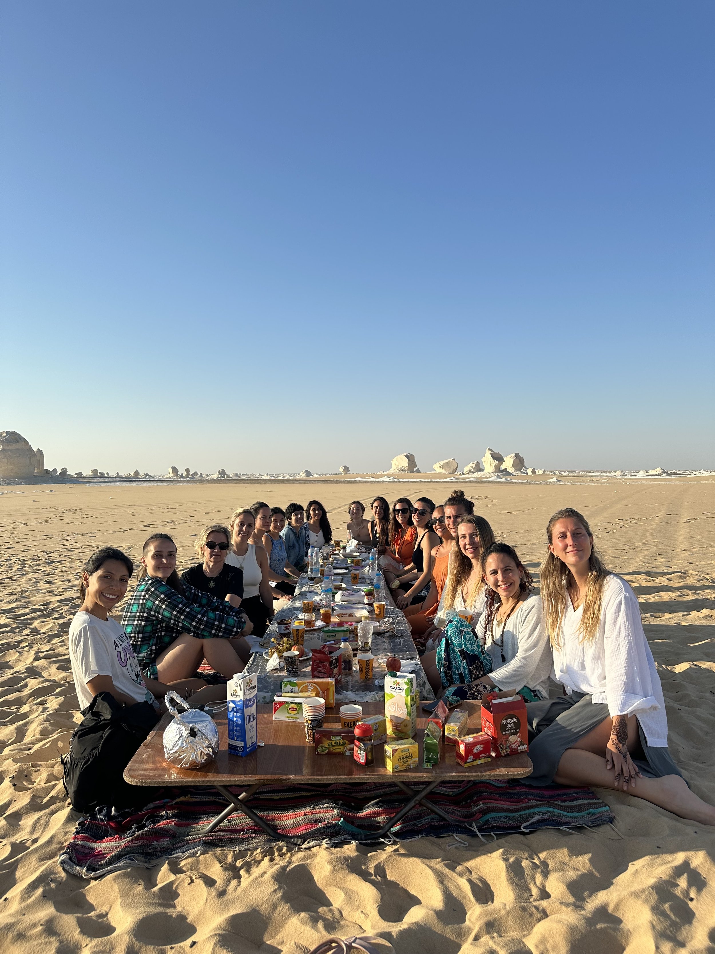 Grupo de personas disfrutando de un picnic en la playa, sentados en una mesa en la arena con comida y bebidas, durante un día soleado.