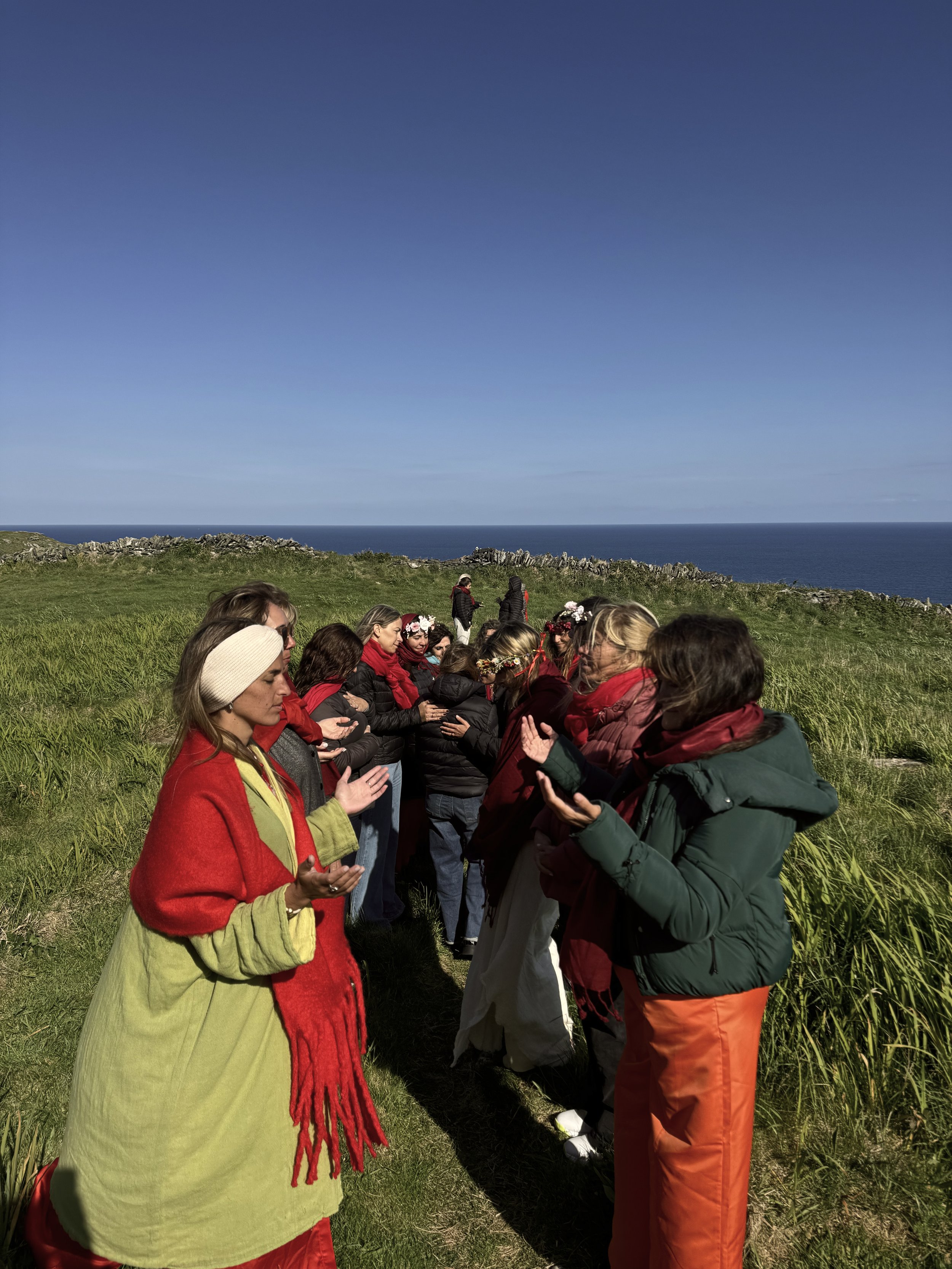 Grupo de personas en un campo de hierba realizando una ceremonia o rezo, con el mar de fondo bajo un cielo despejado.