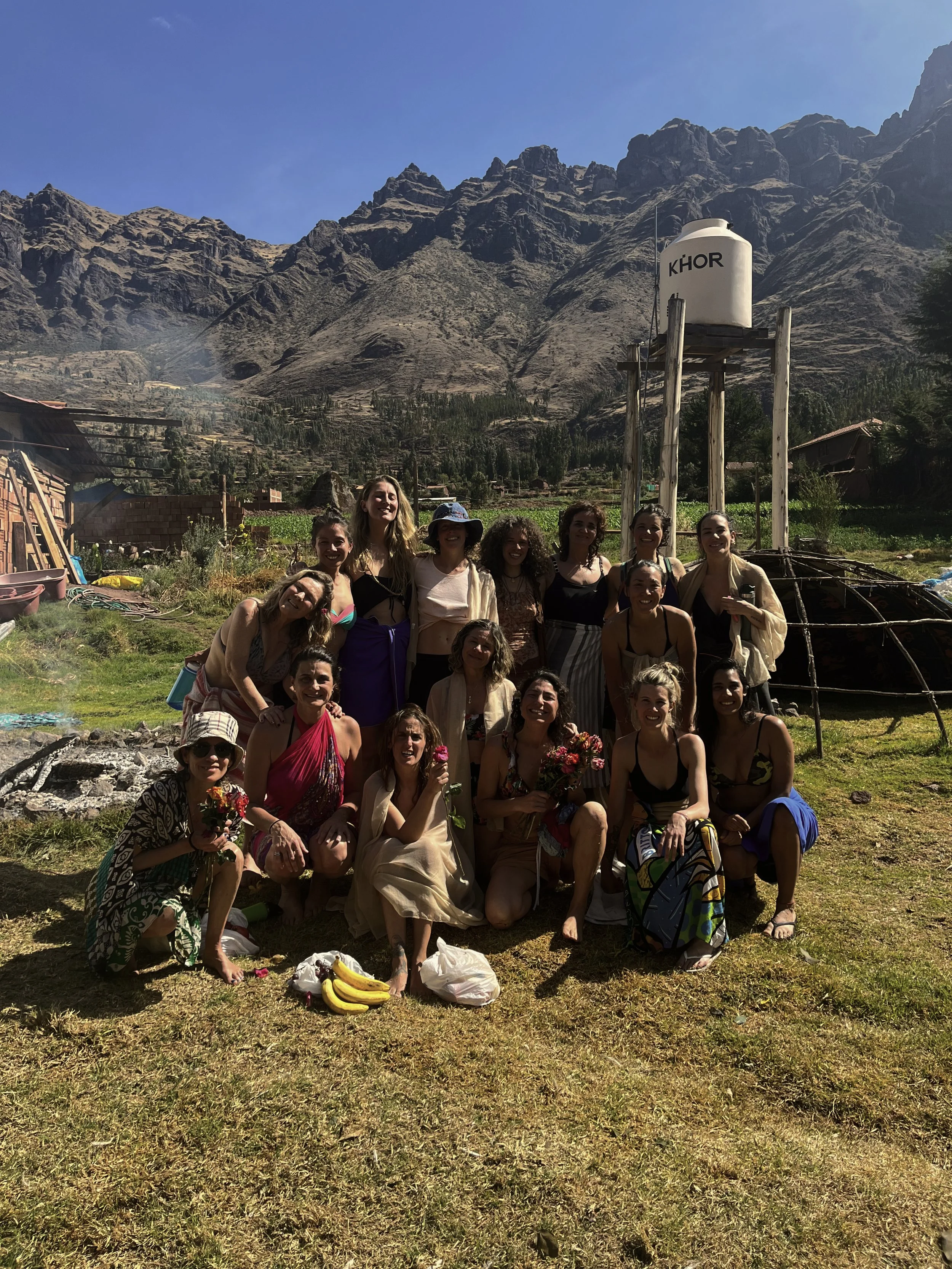 Grupo de mujeres y niñas sonriendo y posando en un campo verde en las montañas con un tanque de agua marcado con 'KHOR' en el fondo.