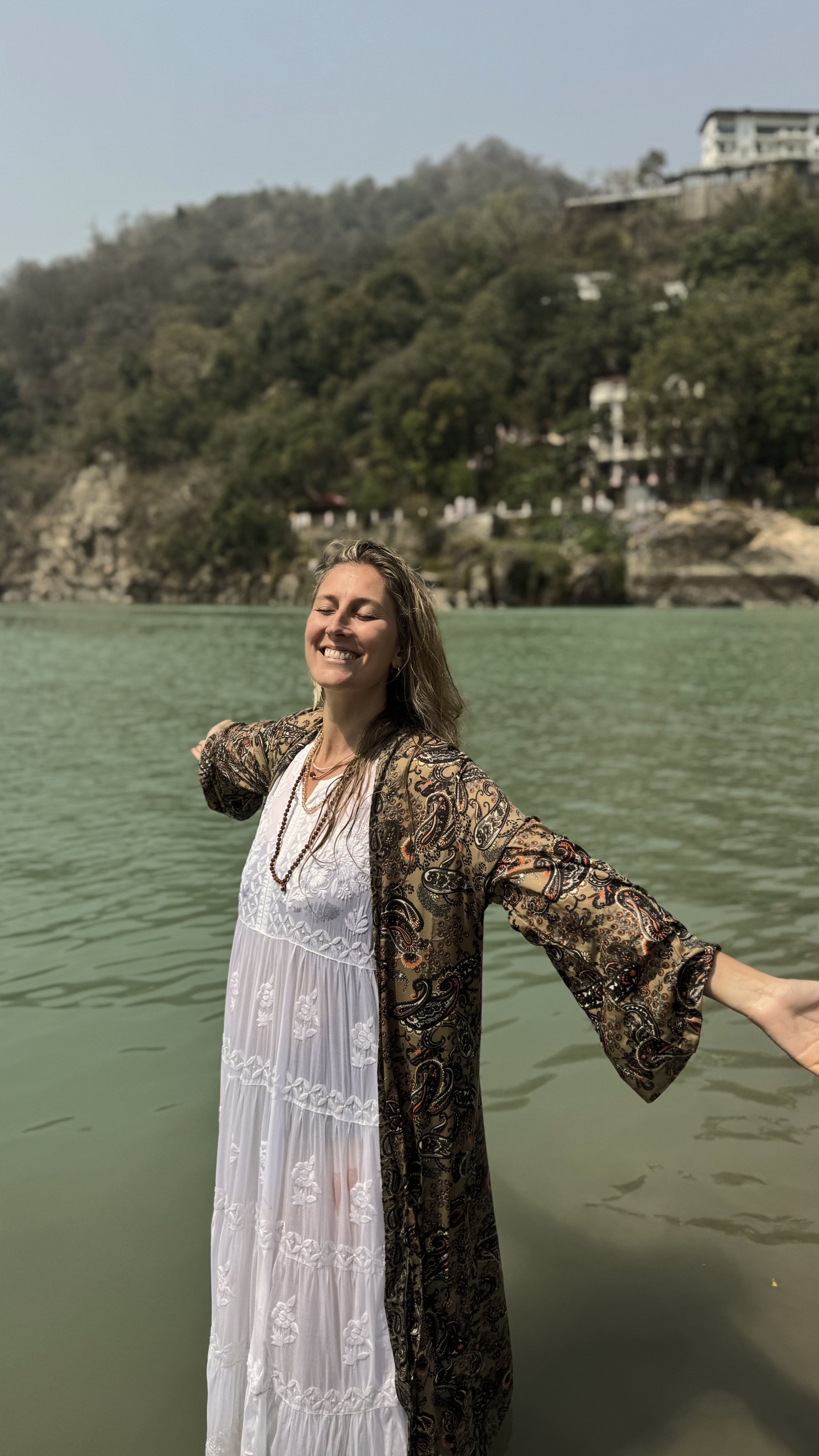 Mujer sonriendo con brazos abiertos en un lago con colinas y edificios en el fondo.