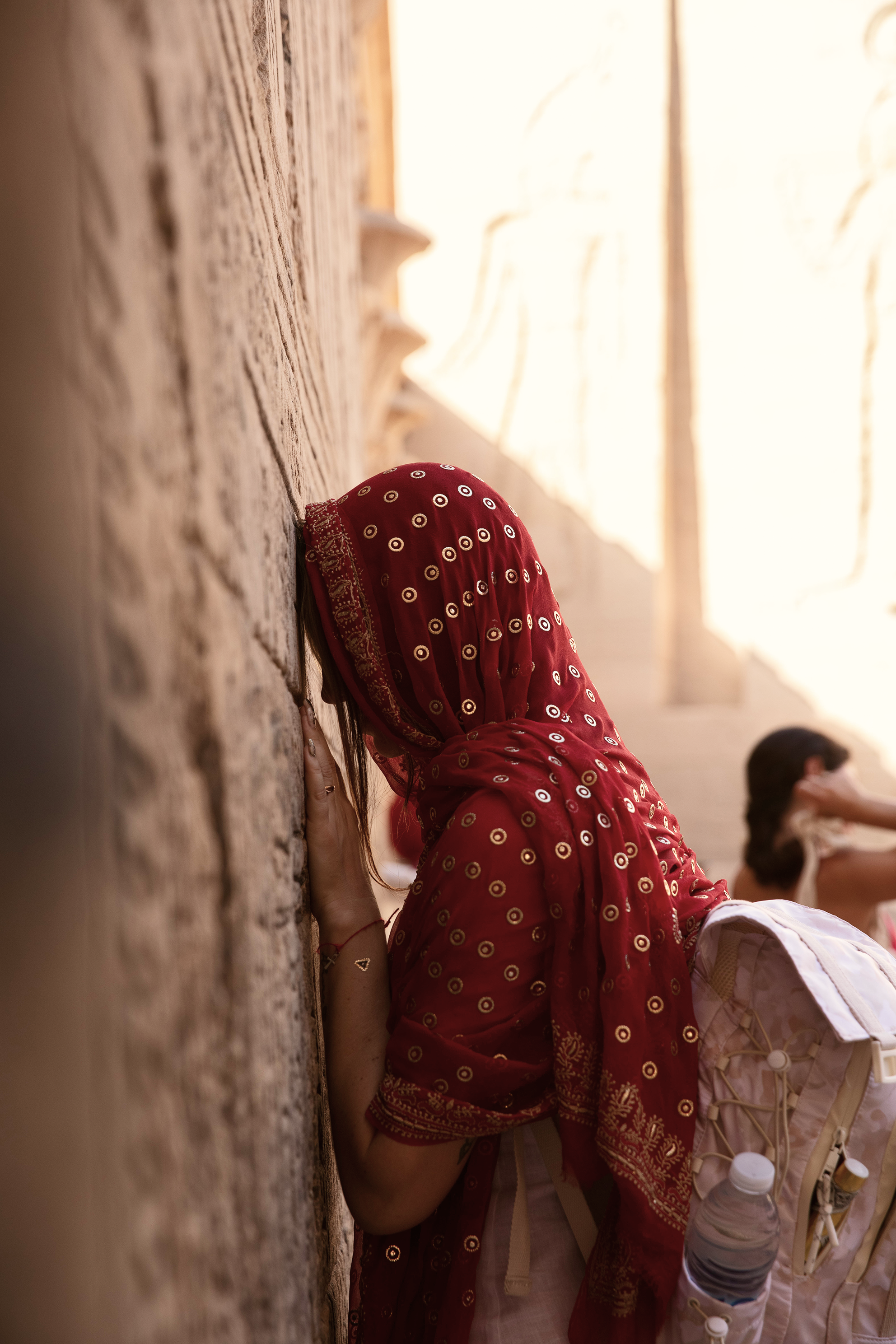 Una mujer con camino en su rostro, con tocado rojo y mochila con botella de agua, apoyada contra una pared de piedra en una escalinata de piedra y estuco, en un lugar donde parece un sitio religioso o histórico.